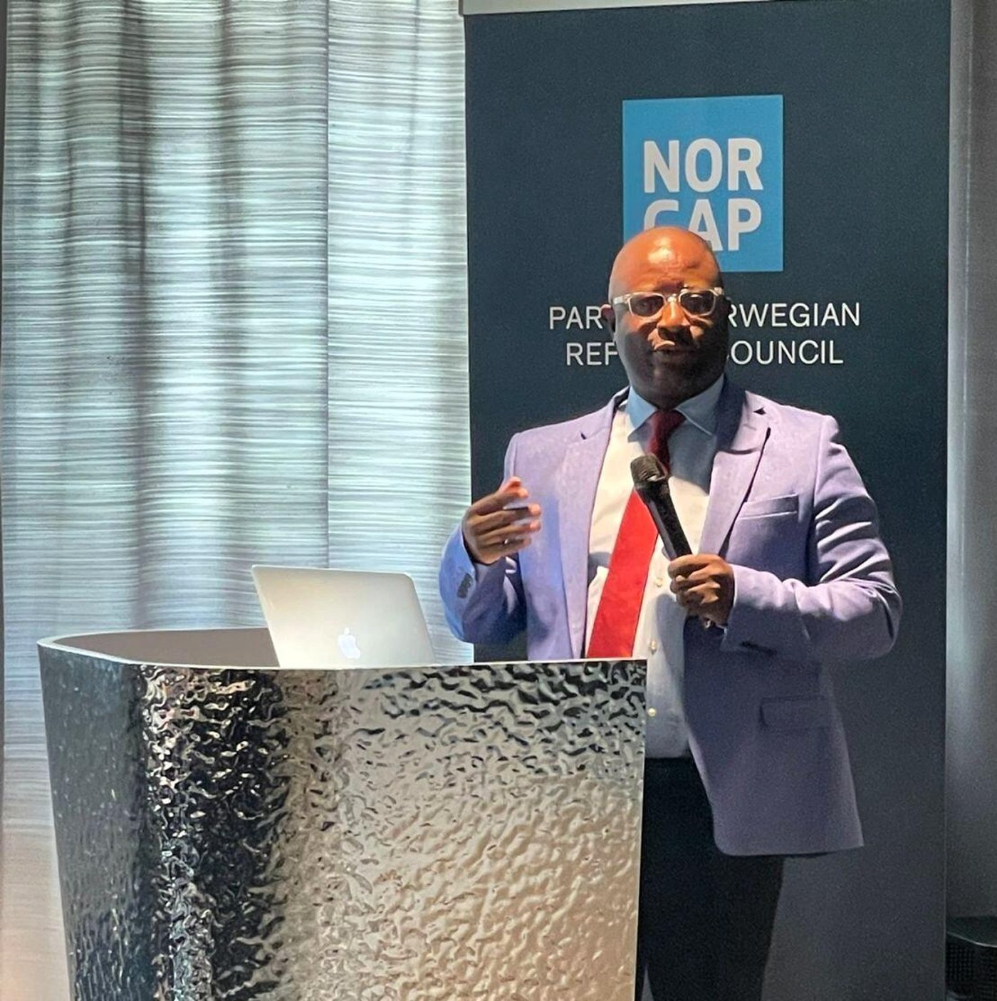 African man with microphone speaking at an event. NORCAP logo in the background. 