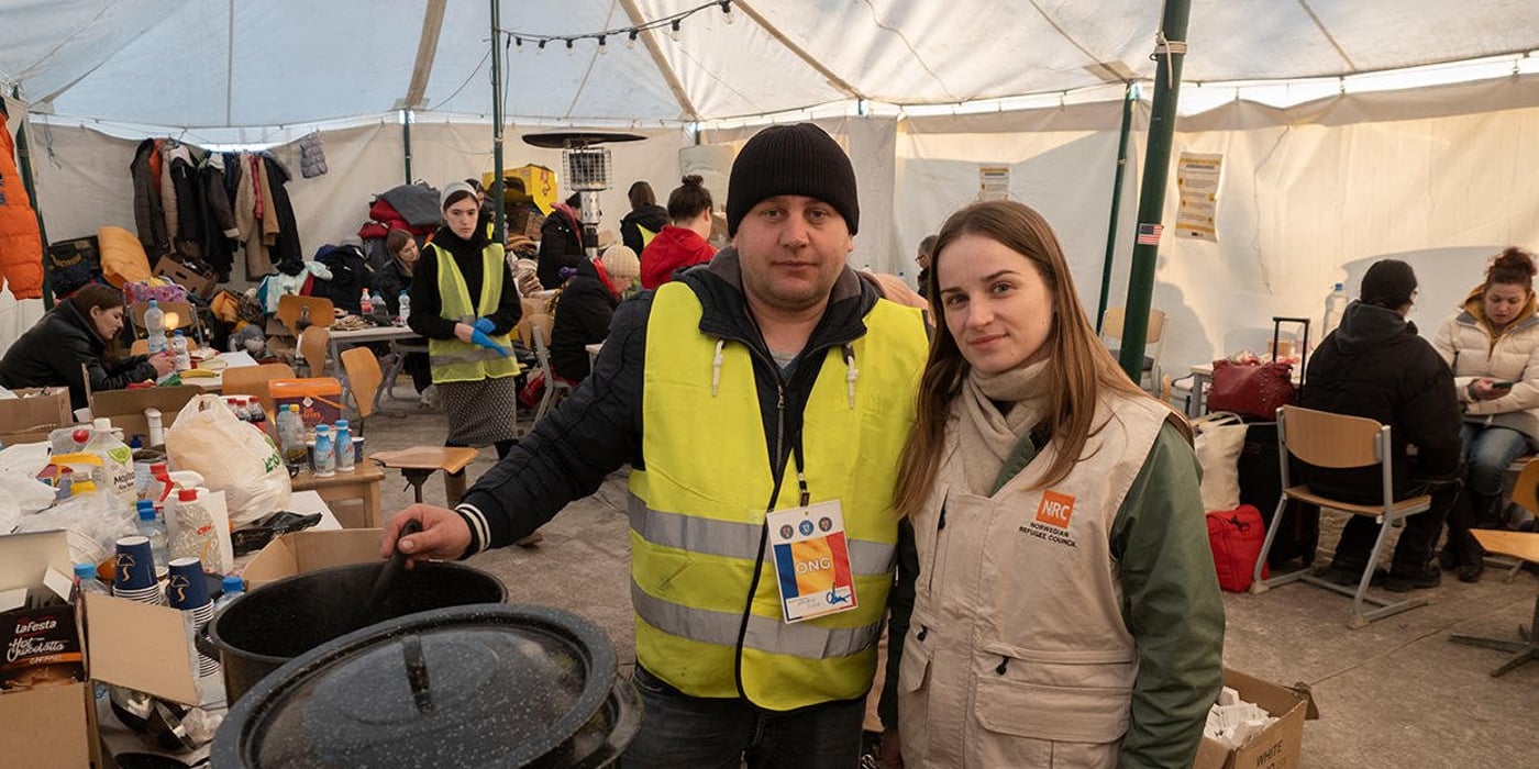A man in a reflective vest and a woman in a beige vest is standing next to each other. In front of them are two big soup terrines. They are standing in a large tent.