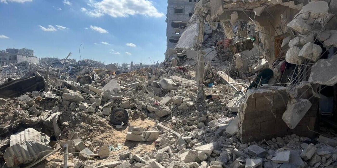 Sönderbombade byggnader och stora mängder bråte i ett bostadsområde i Gaza, med blå himmel i bakgrunden.