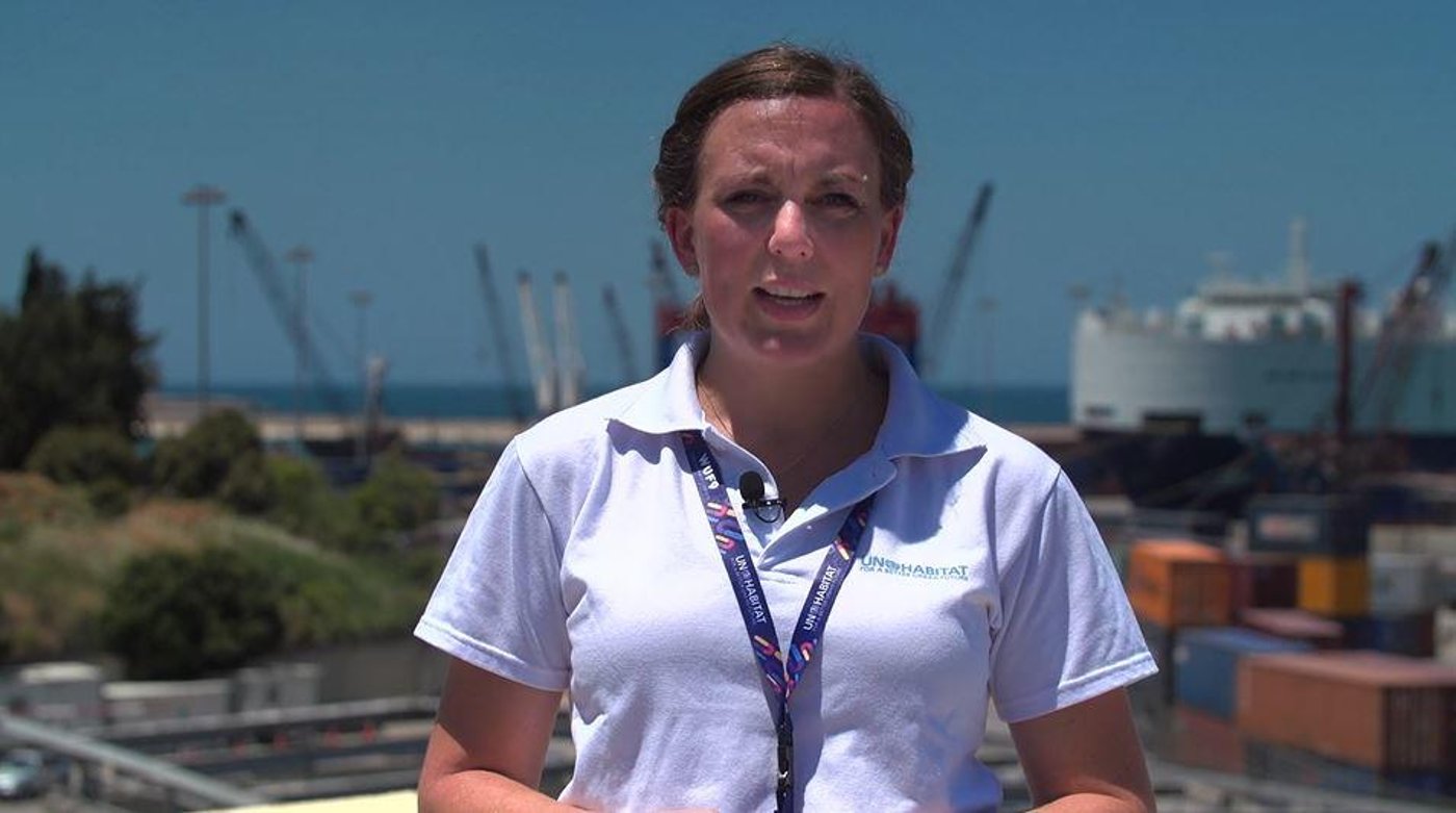 Woman standing in the middle of the photo, port structures behind her, speaking directly into the camera.