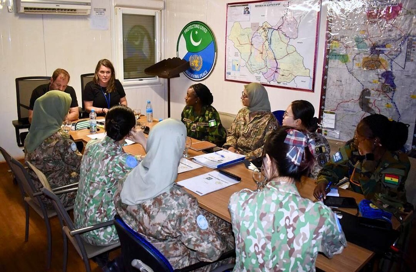 10 people gathered around a table, the majority young women in military uniform, talking to two people in black at the end of the table. Big maps on the wall behind them. 