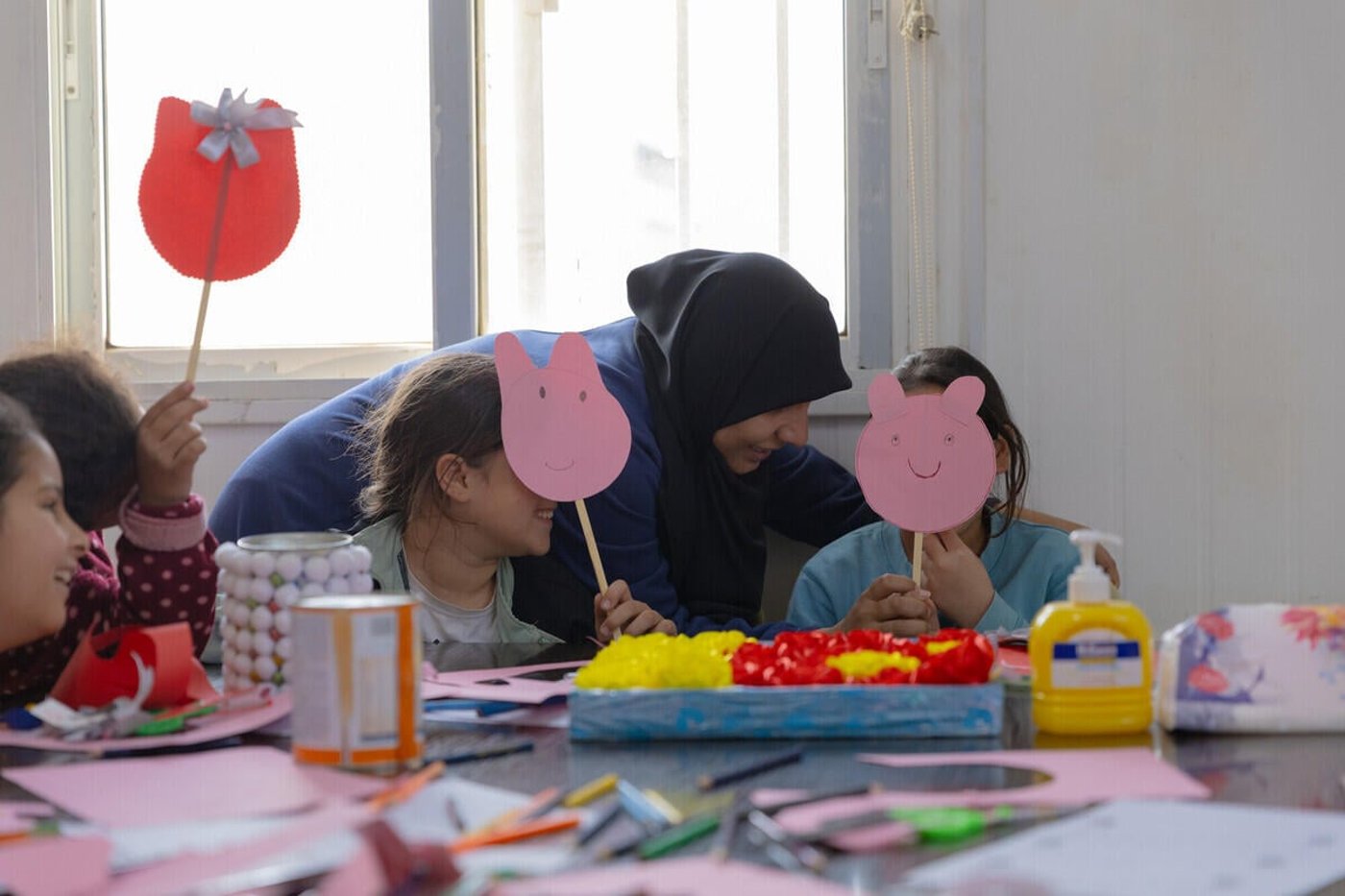 Two children hiding behind pink masks they have made. A teacher is leaning towards them.