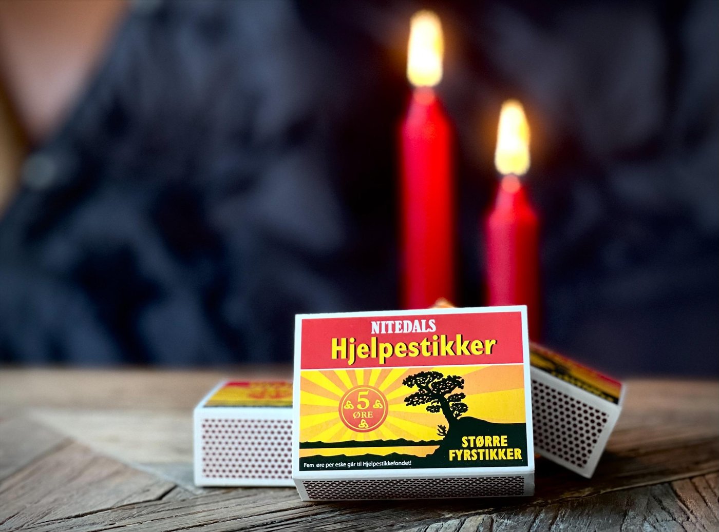 Three boxes of Nittedals hjelpestikker (matches) is lying on a wooden table. Behind them are two lighted red candles. The background is dark and blurry.