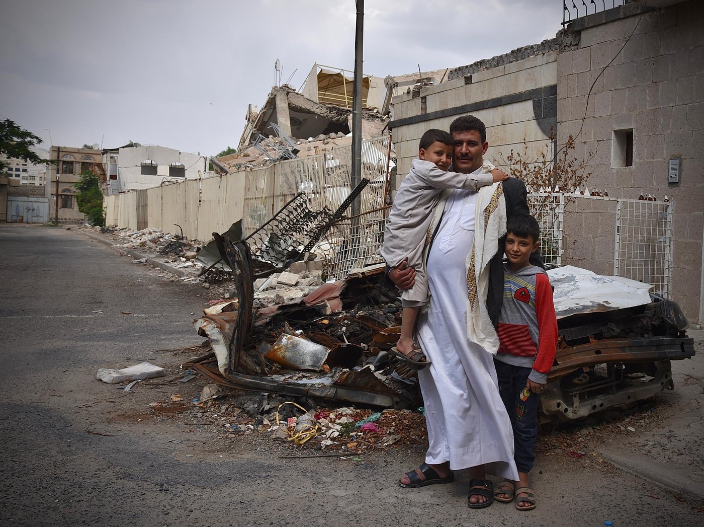 From left: Children Khalid (6), father Abdulrahman and Saif (11).
“God forbid, I could have died that evening.”
Abdulrahman is the father of three young children and works as a compound guard for a local organisation in the Haddah neighbourhood in Sana’a. The family live right next to the compound in a guard’s house.
On the evening of 5 June at around 9pm during Ramadan, the family were inside their house resting. Abdulrahman sent his youngest son across the road to bring water. Usually, Abdulrahman himself would be outside wandering around the area, but this evening he decided to stay in. Only minutes after his son arrived back home with the water the family heard a loud explosion and felt their house violently shake. It was the sound of a bomb that had just been dropped within metres of their home by a Saudi-led coalition aircraft.
“The first bomb they dropped did not explode. Two or three minutes later, the second bomb was dropped and we got very afraid and tried to secure ourselves inside the house,” Abdulrahman said.
Abdulrahman moved towards the window to try to see if he could assess the damage.
“That’s when the third bomb was dropped. I was thrown back against the wall as the window of our house exploded. We ran down to the basement. My children were screaming for their lives at this point; it felt like complete horror.”
Abdulrahman got fragments of the window glass in his leg. As the family reached the basement, a fourth bomb exploded and caused the two-story concrete structure next door to implode. The family that live their had travelled away in advance of the Eid holiday.
“We never expected something like this to happen so close to our home. My youngest son still runs to a corner and hides whenever he hears planes hovering above us. We had to take them away from this area and our house for a while to allow them time to process.”
The attack injured seven civilians including four children. It also damaged several houses, including a neighbouring house rented by NRC for our expatriate staff, with staff inside at the time.
Photo: Becky Bakr Abdulla/NRC