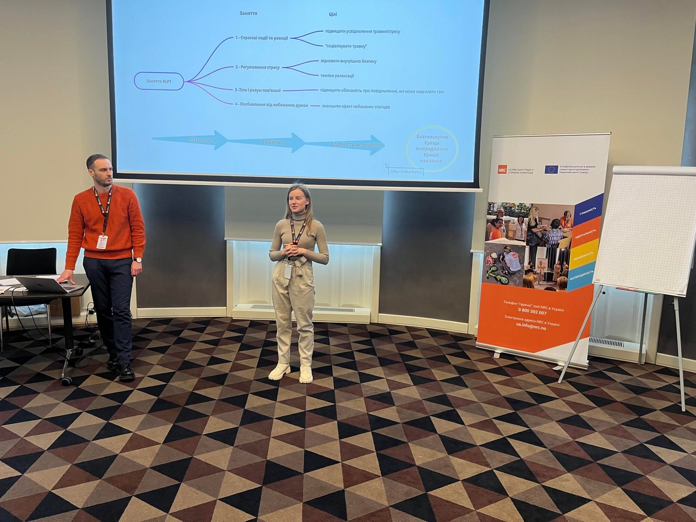 NRC staff members Mariia and Anatolii are standing in front of a screen in a conference room. The screen displays a presentation.