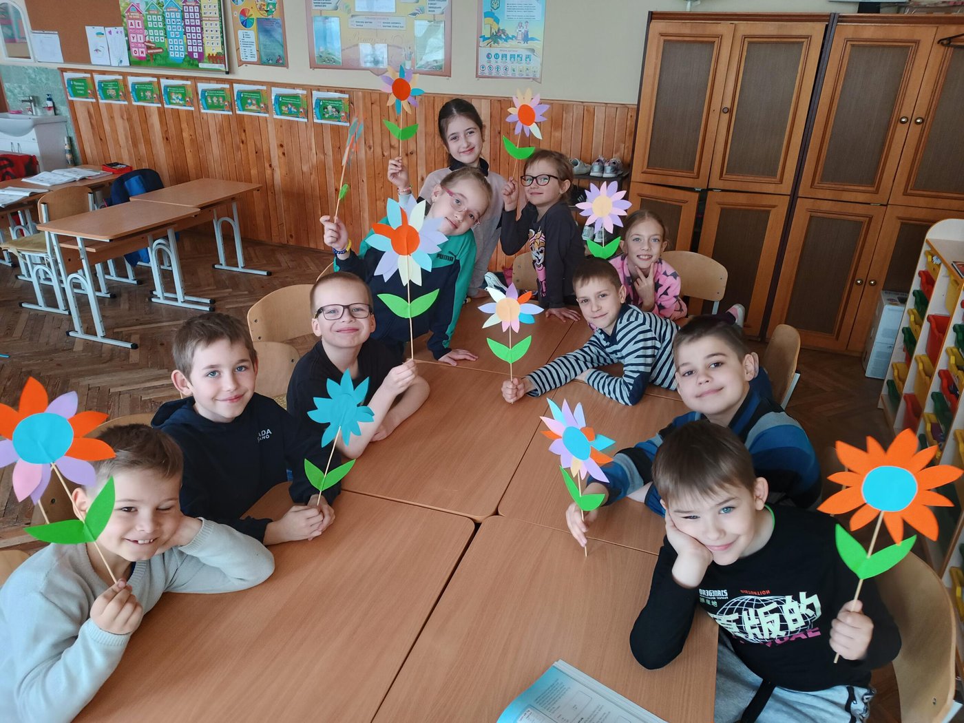 A group of children sitting around tables grouped together to form a rectangle. They are all holding paper flowers of different colours. 