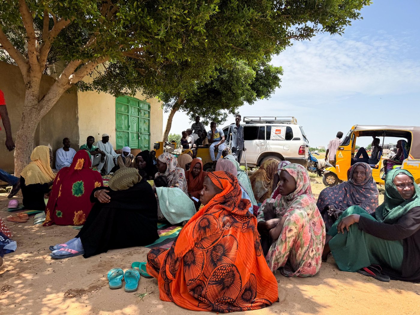 Sudanese women sitting in a group under a tree. Backs to camera and most faces turned towards the left side of the photo. Some cars are in the background, one with an NRC logo. 