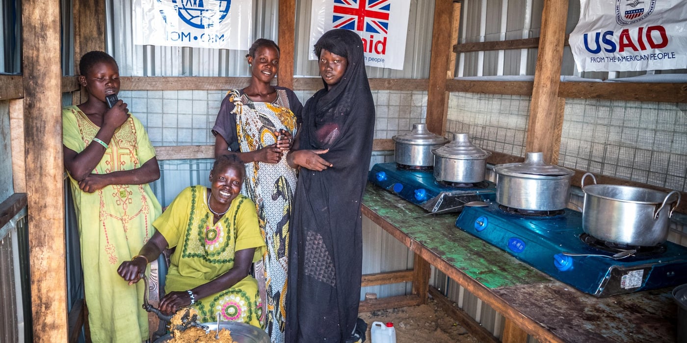 Four women standing in a communal kitchen, one grating food while others mind the pots and pans on gas stoves. (Photo: Iban Colon/NORCAP)