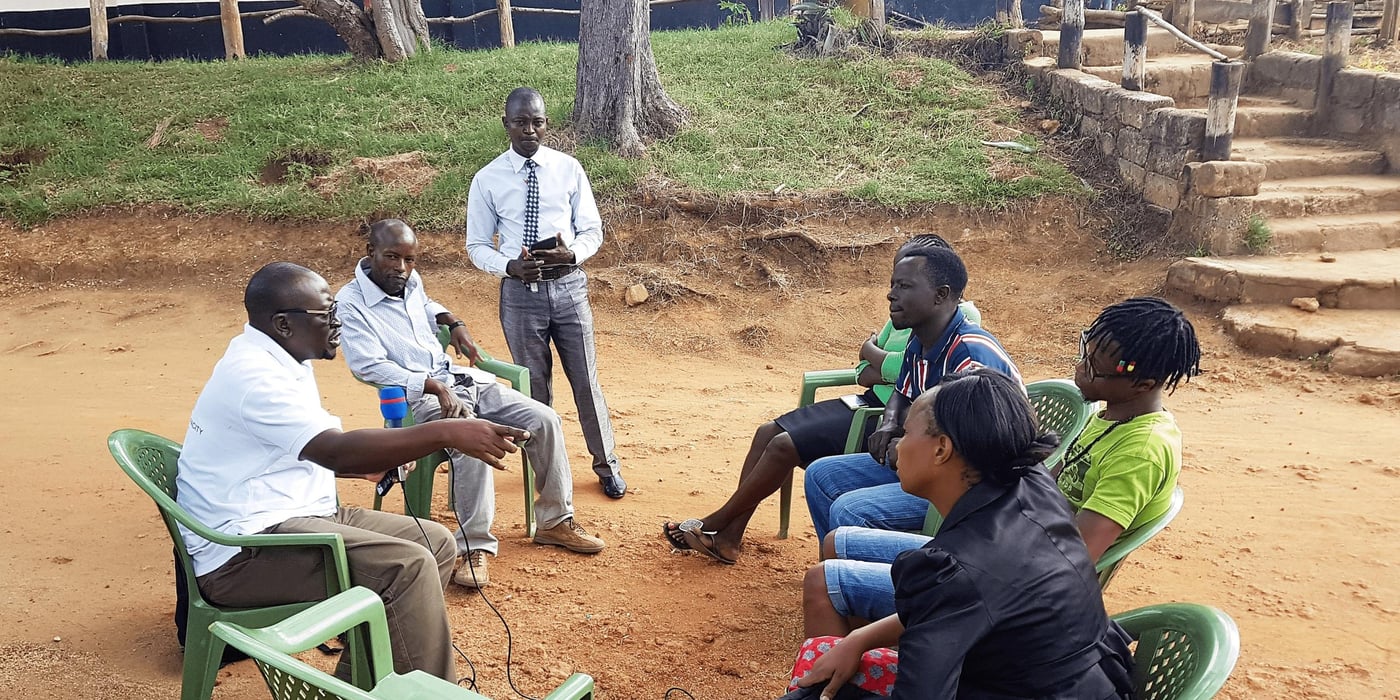One man interviewing four community members while two other  men sit in the background observing. 