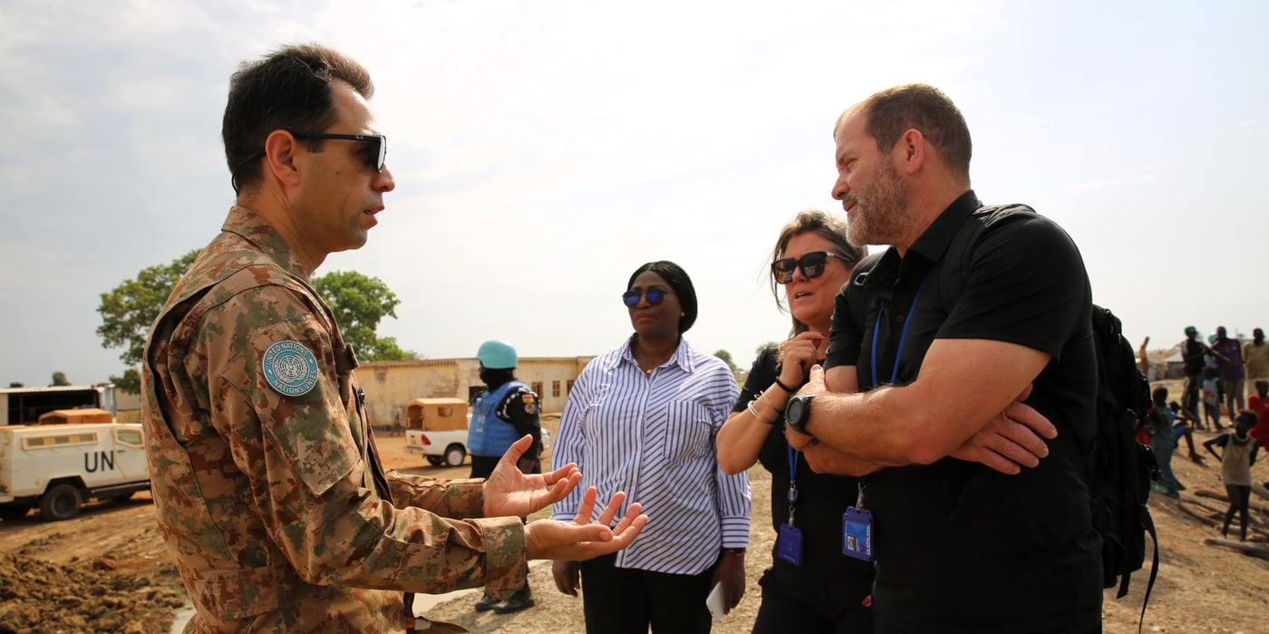 A man in military uniform talking to two people in black clothing in a rural setting. In the background, a United Nations vehicle, a UN soldier and people from the local community.