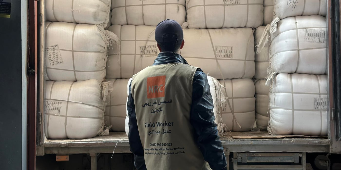En man står bakom en lastbil lastad med stora vita säckar, staplade tätt intill varandra. Han bär en väst märkt med Norwegian Refugee Council och texten – Field Worker – och ser ut att inspektera eller förbereda lossning av humanitärt bistånd.
