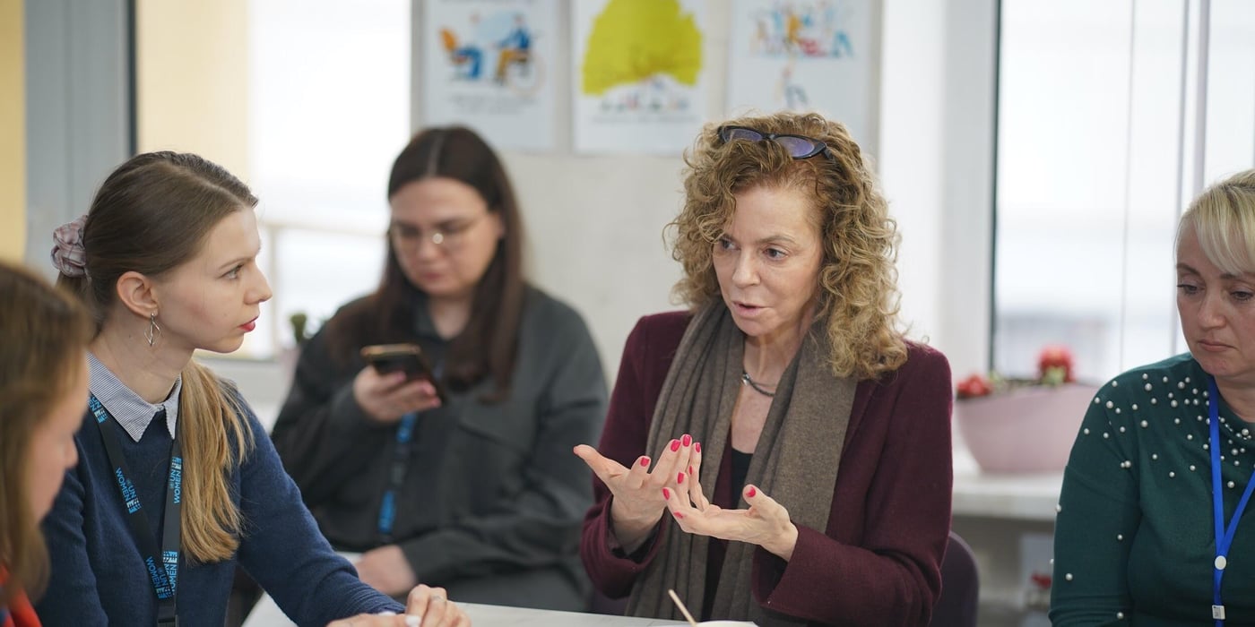 Young and middle-aged women sitting around a table, engaging in conversation. One woman sitting in the background, checking her phone. 
