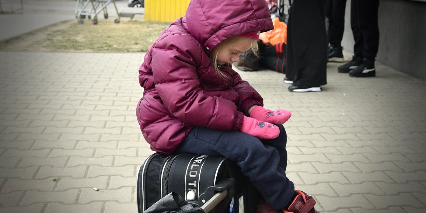 A young Ukrainian girl is sitting on a suitcase, looking down at her hands. She is wearing a pink coat and pink mittens.