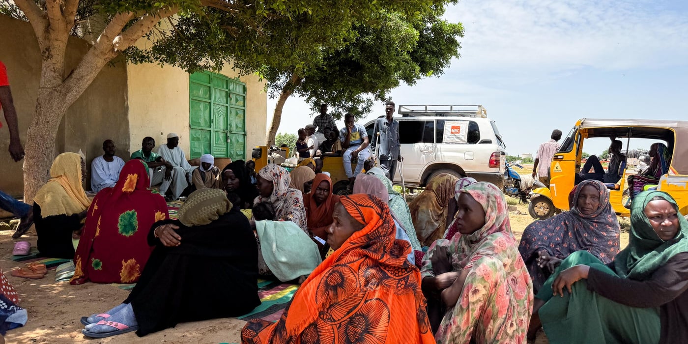 Sudanese women sitting in a group under a tree. Backs to camera and most faces turned towards the left side of the photo. Some cars are in the background, one with an NRC logo. 
