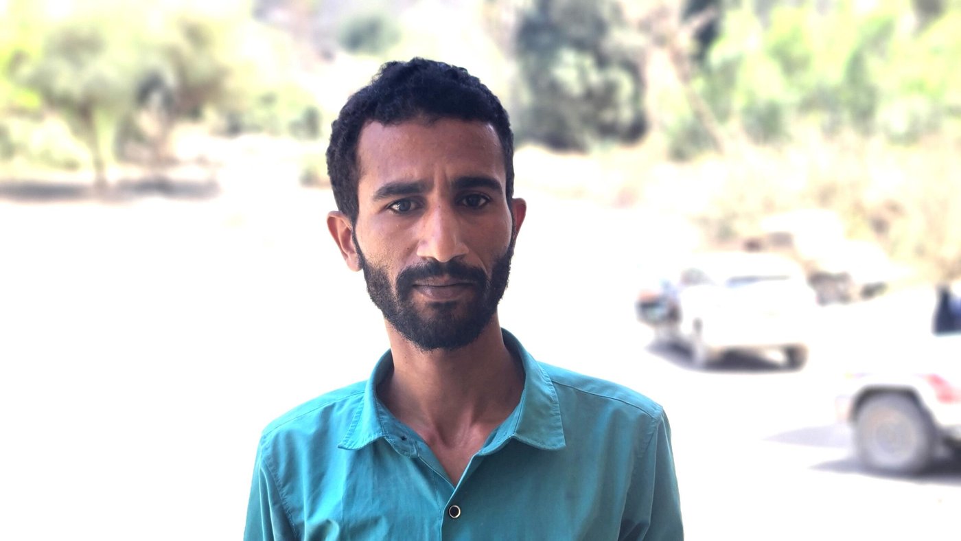 Portrait of a Yemeni man. He is wearing a blue shirt. Behind him are a few cars and trees.