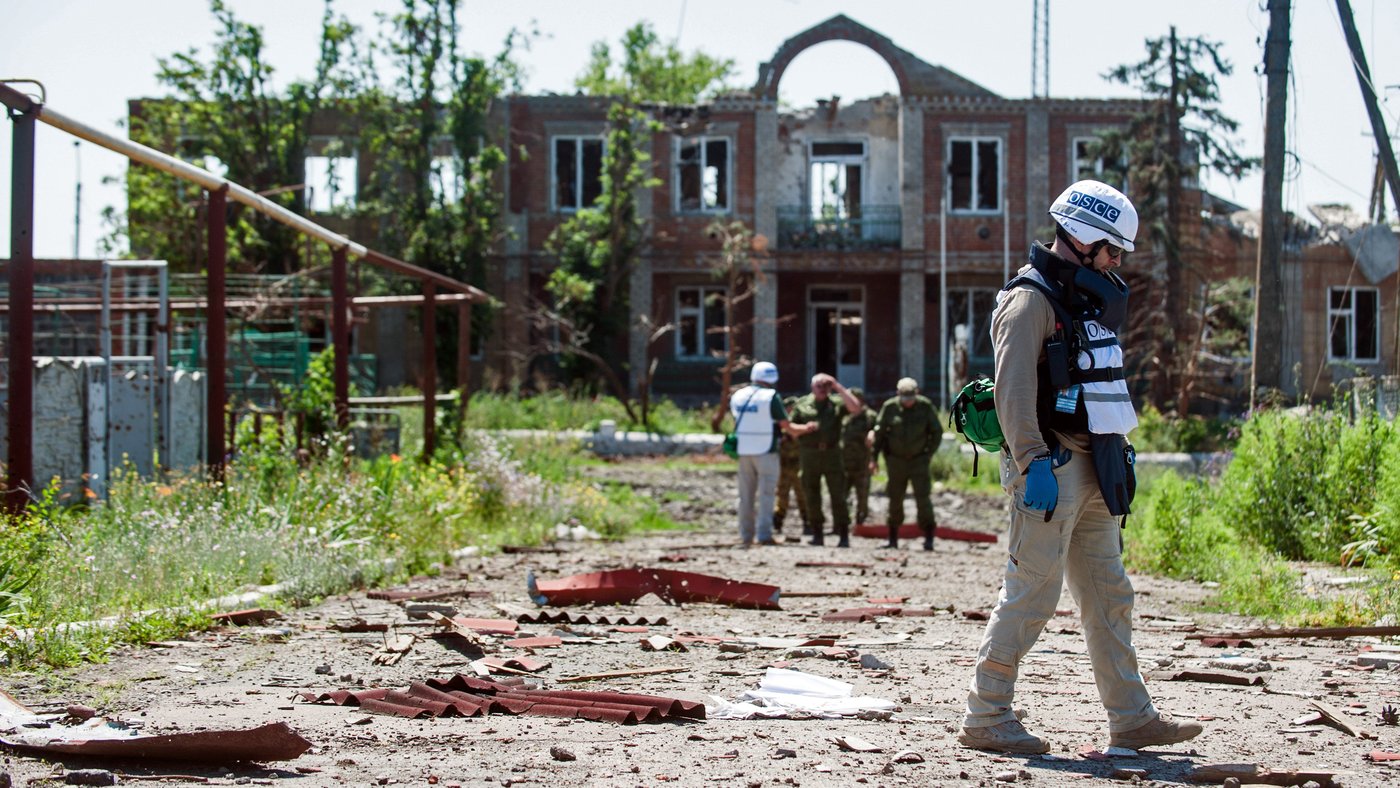 A man in helmet and kevlar vest observing the ground scattered with debris. In the background, a group of men are talking, some in the same protection as the man in the foreground. The group is standing in front of a bombed out building with no roof and windows. 