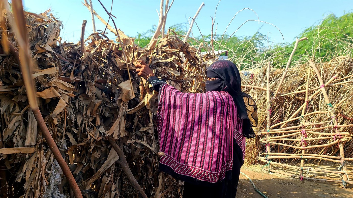 Fatima holding a banana leaf against her shelter, which is made entirely out of the same leaves.