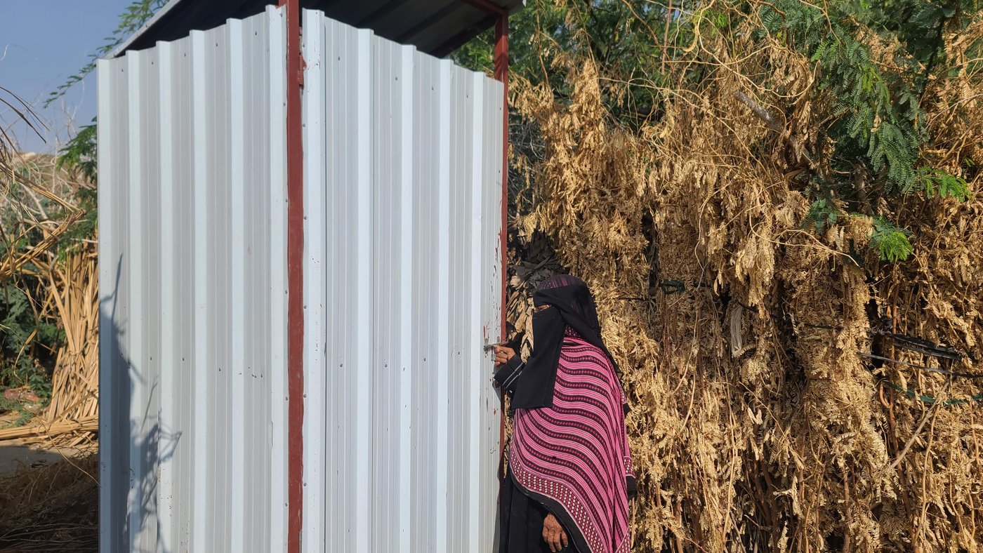 Fatima stands by her latrine, which is made out of metal sheets. There are trees in the background.
