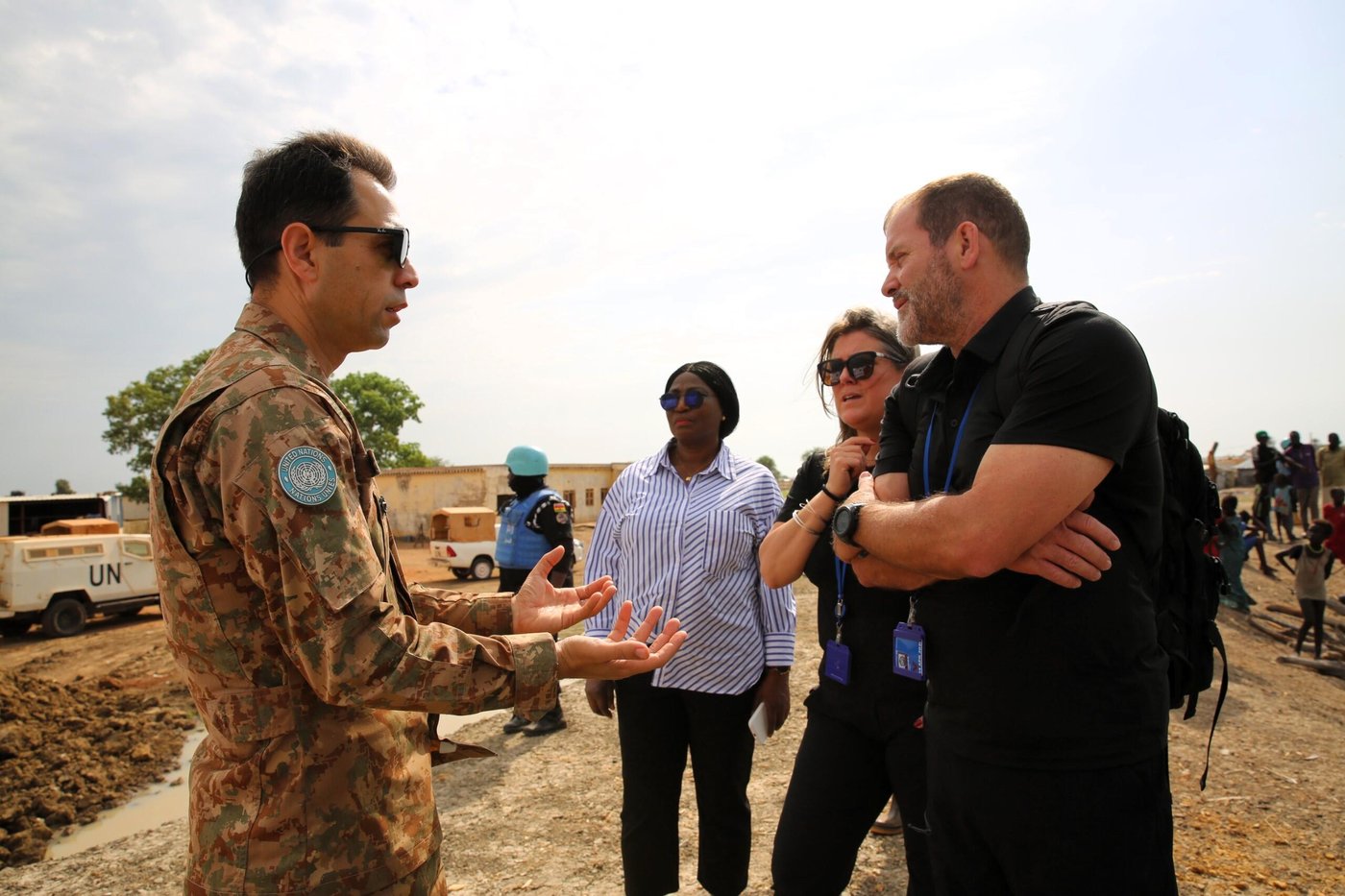 A man in military uniform talking to two people in black clothing in a rural setting. In the background, a United Nations vehicle, a UN soldier and people from the local community. 