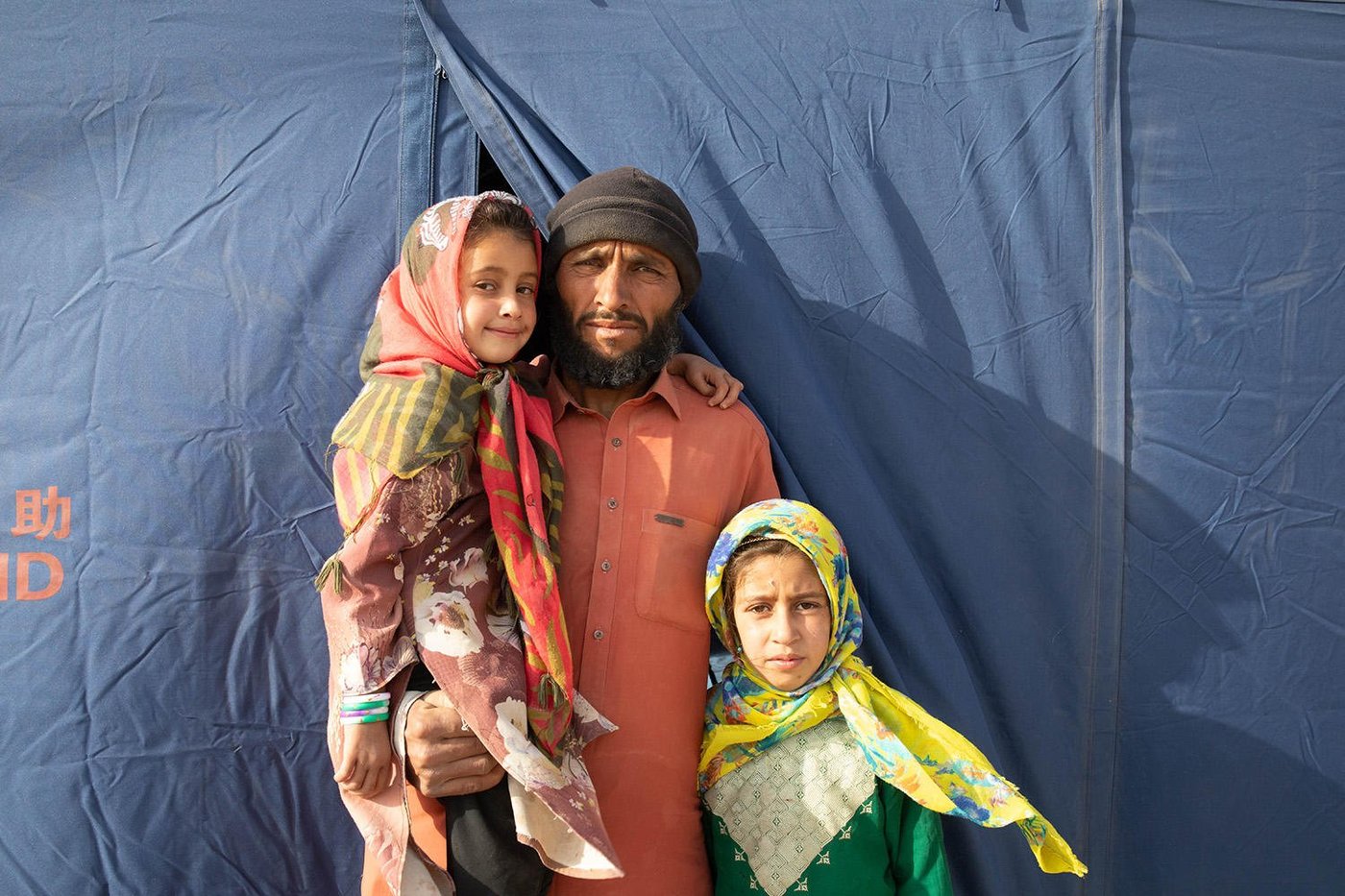 A man with two of his children are standing outside the tent they are living in. He is holding one daughter, while the other is standing next to him.