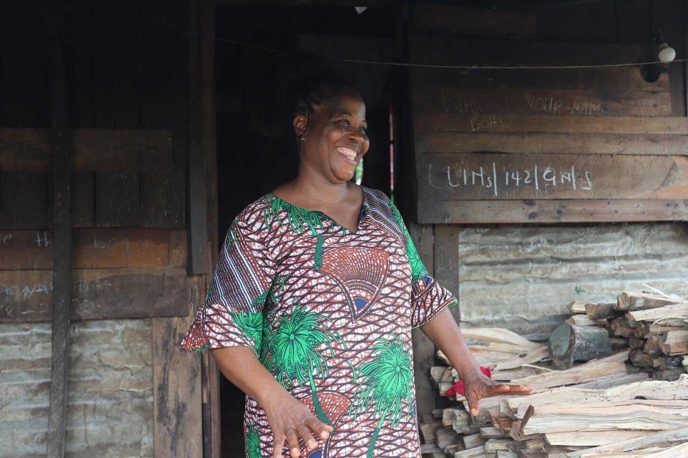 Vivian is standing by a table piled with thin wooden logs. She is smiling broadly with one hand resting on the pile of wood. Behind her is a wooden wall with chalk writing.