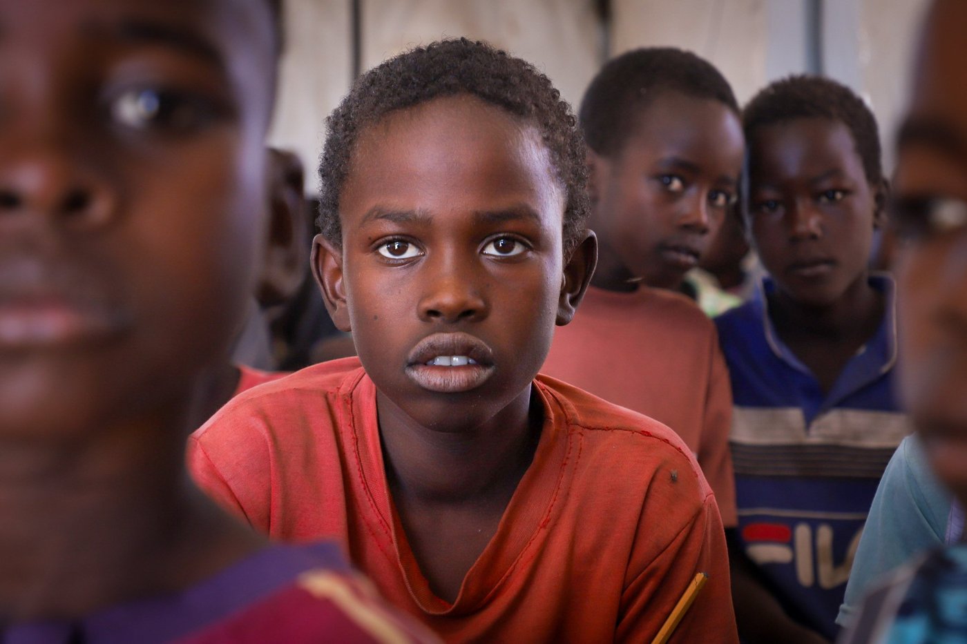 En pojke i röd tröja sitter i ett klassrum med andra barn och tittar in i kameran.