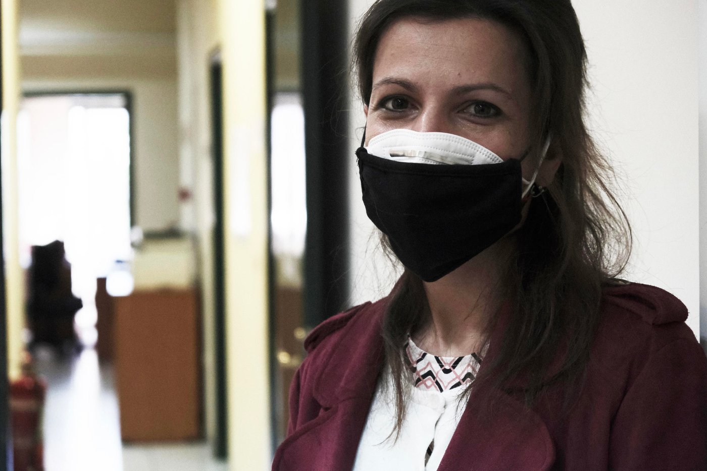 Profile shot of a woman wearing two face masks, looking straight into the camera, from inside an office building.