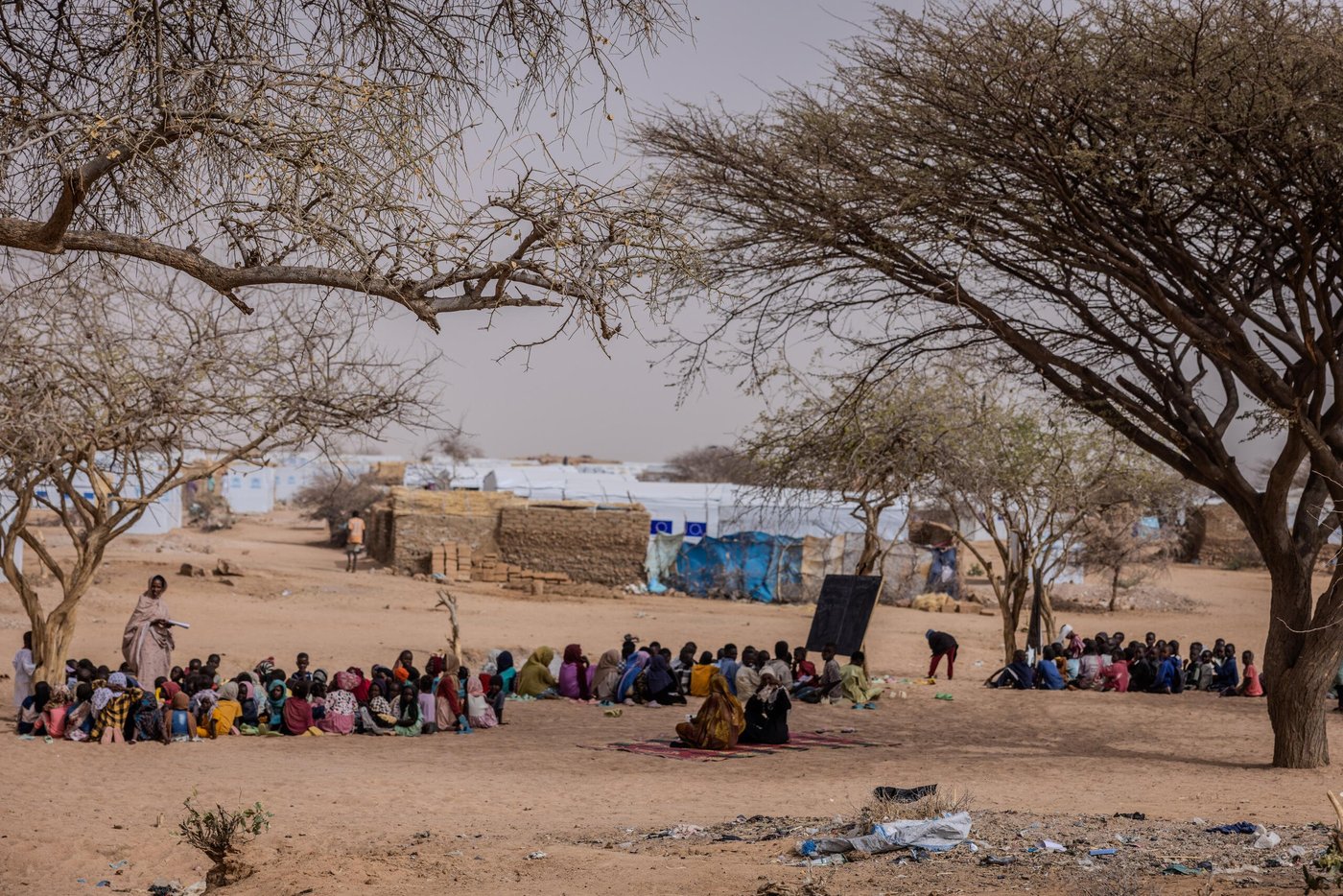 En grupp kvinnor och barn sitter samlade i skuggan av några träd i ett torrt och dammigt landskap. I bakgrunden syns enkla skydd byggda av lera och presenningar i ett flyktingläger.