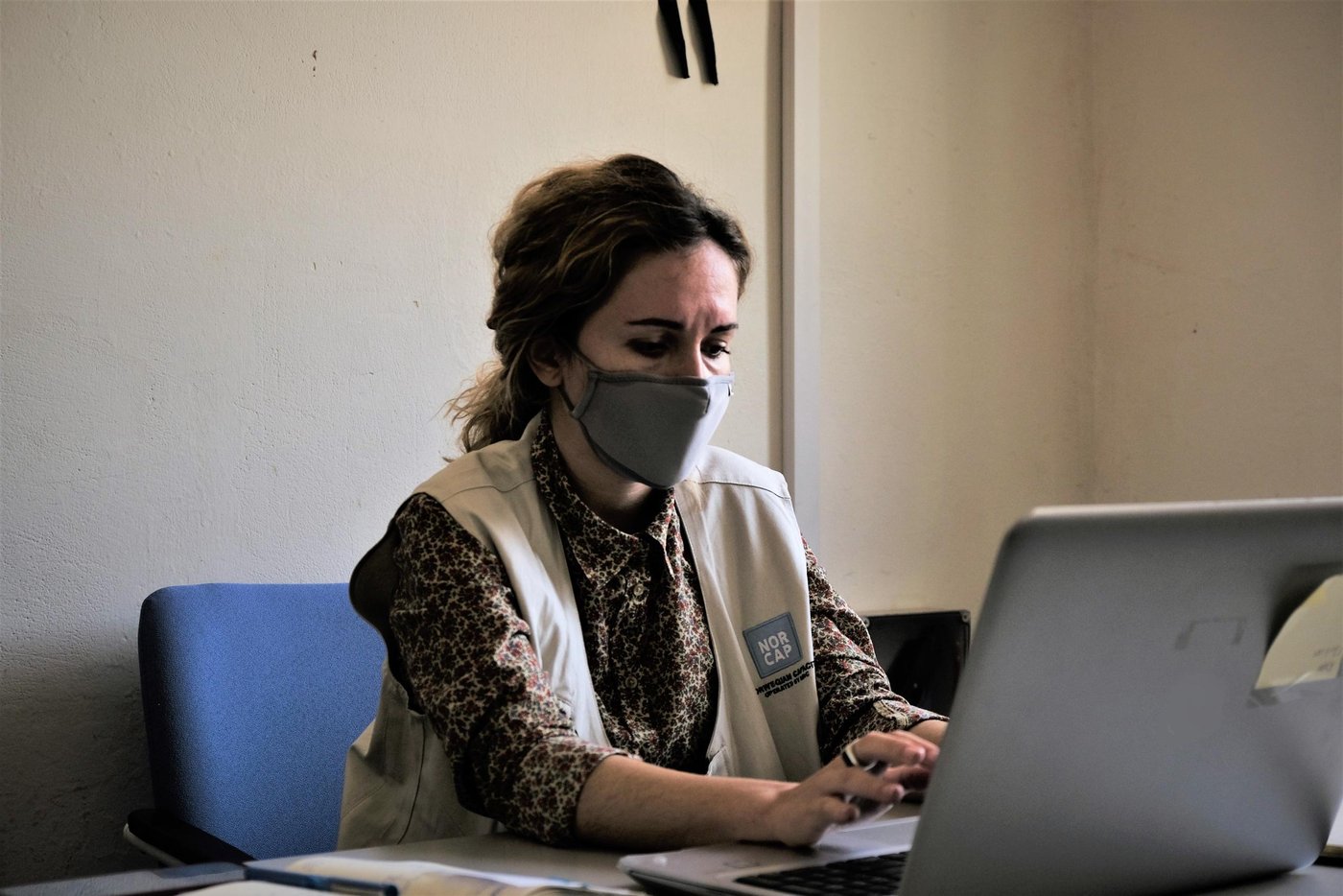 Woman wearing a face mask sitting in front of a computer screen, writing on a laptop.