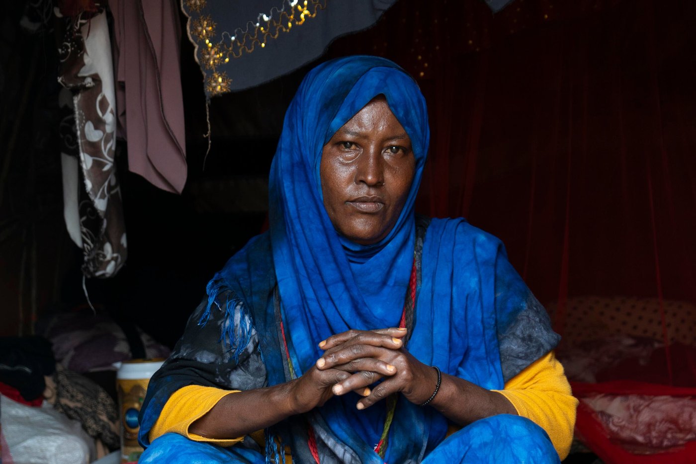 En kvinna sitter inne i ett dunkelt tält och ser rakt in i kameran. Hon bär en klarblå hijab som täcker hår och axlar, och en gul tröja syns under tyget. Händerna är sammanflätade framför henne.