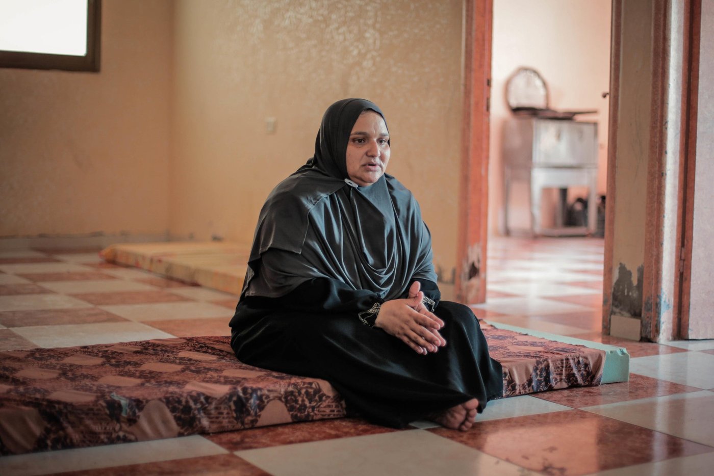 Jumana Murad, 47, in her bare rented apartment.
In just over seven years, Jumana, who lives in Al Shati refugee camp near Gaza City, lost four of her children.
Juliana died at age 17 in 2012 after she accidentally took the wrong medication for a severe stomach ache. Just over a year later, Lina, 14 at the time, died when she fell from the fourth floor of a building. Jumana says her son, Qusai, inhaled smoke from Israeli shells fired during the first major round of hostilities, which ran from December 2008 to January 2009, and developed respiratory problems that contributed to his death in 2014 at age 6. 
This year, Jumana grieved the loss of her 19-year-old daughter, Jasmin, who died from complications of kidney failure.
Medical bills have left the family in debt. 
All told, the family owes 2,900 NIS (US$825) for groceries, prescriptions, and rent. Unpaid promissory notes to landlords have landed Jumana’s husband, Adel, a tailor by profession, in prison since July 2018. After losing his job at a factory in Israel, he worked as a cleaner in Gaza until health problems resulted in too many absences and he was let go. 
According to the Ministry of Public Works and Housing, some 2,000 households faced the risk of eviction in 2018 over inability to pay rent. As of August this year, the Ministry of Social Development pegged the number of families under eviction risk at 9,356. 
Evictions have become routine for Jumana and her family over the past two years. They have found themselves often sleeping on the street or at the port several times.
“When we were in the tent in the street, we felt very embarrassed whenever people walked past us,” said Jumana. “We tried to cover ourselves with plastic and carton sheets. We did not have water, so we could not shower, and our poor personal hygiene caused us skin problems.”
While in the tent and on the street, the family relied on the owner of a nearby restaurant and goodwill from other people for food. Jumana’s boys also scavenged the local market for leftovers near spoiled vegetables to complement their diet of bread and tea.
All five of Jumana’s surviving children dropped out of school because of the number of times they have been forced to move after getting evicted from their home, as well as lack of funds for school fees, uniforms, and transportation. Ali and Mohammad, 15, completed first grade only; their siblings Jenin, 17, and Saed, 16, made it to second grade, and Diana graduated from fifth grade.
The family receives 1,600 NIS (US$453) in cash assistance from the Ministry of Social Development every three months, which are often delayed or incomplete, and UNRWA food vouchers, but often uses them to repay grocery debts. In fact, more than half of the households surveyed by NRC resorted to selling the assistance received.
Since May, Jumana’s family has received cash-for-rent from NRC, as part of a project funded by the Norwegian Ministry of Foreign Affairs to support 280 vulnerable households to stave off the threat of eviction.
“We are terrified of living on the street after the cash-for-rent assistance from NRC finishes,” said Jumana. “We always think about what we are going to do after the end of the project. I dream about having one room for myself one day.”
The loss of the children, “one after the other," as Jumana says, has taken its toll, physically and psychologically, on the surviving family members. Adel, and Jumana’s oldest son, Ali, both have psychological problems and require expensive prescriptions.  
Ali, was only 15 when he had witnessed his sister plummet to her death. A four-month stint in a psychiatric hospital barely scratched the surface, and he increasingly turned to drugs and crime. Now 21, he has been serving a seven-year sentence for numerous robbery convictions since 2017.
While the prison has granted Ali furlough on weekends, he has used the time, together with his family, to participate in the Great March of Return mass protests on Fridays along Gaza’s perimeter fence with Israel. Without hope or prospects for the future, Jumana says they take advantage of the free transportation to attend the demonstrations for a sense of stress release from their daily lives.
Unfortunately, Ali, his sister and father were unable to escape injury. 
Ali was shot twice in the leg, in separate incidents in 2018, sustaining serious nerve and artery damage. His latest injury occurred in January this year, when a rubber-coated steel bullet fractured his nose. 
Diana, 18, sustained shrapnel wounds to her left arm and thigh in 2018. In March this year, a tear gas canister struck her in the right shoulder. While Diana’s father looked for her amid the crowds, he fell to the ground with a bullet wound to the leg. Complications arising from diabetes and kidney problems have stalled his recovery.
Photo taken on 21 August 2019
Photo: Ahmed Mashharawi/NRC