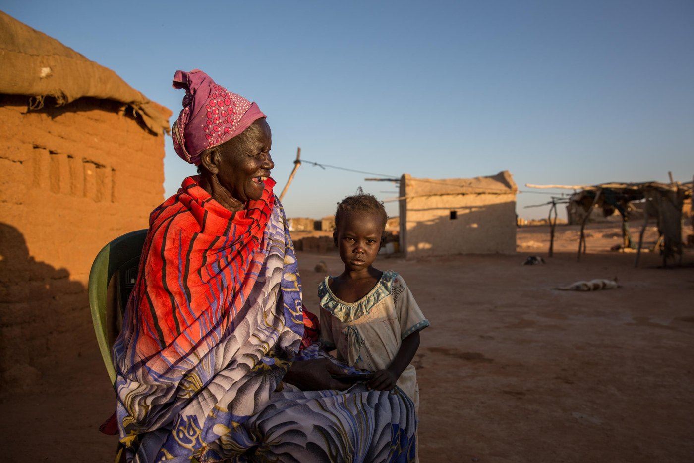 Apuk, 70 year old grandmother in her home.
Full story:
Anglo Woku never imagined he would be considered a refugee in Sudan in his twilight years. Especially since he left the Southern part of the country for a better life almost 50 years ago.
“I escaped in 1969, shortly after the civil war broke out. I ended up in Khartoum. I was working for the military. I was married, had a family and friends. Sudan gave me a good life,” he said.
The conflict between North and South raged on for 22 years until a peace agreement was brokered in 2005. Then in 2011, the people of the South voted for independence and South Sudan, the world’s youngest nation, was born.
This new dawn however caused all sorts of legal uncertainties for the South Sudanese living in Sudan, particularly in the years following independence.
“I lost my Sudanese identity,” Anglo, 78, explained: “I went from being a public servant to a refugee.”
In 2011 following the independence of South Sudan, Sudan passed laws that had a detrimental impact on the identity of people with Southern heritage.
They automatically lost their Sudanese nationality and were now considered South Sudanese. Even their children, despite being born and raised in Sudan, were given a new nationality. Many people had no intention of returning to South Sudan, a country, largely unknown to them and this created a legal dilemma on questions around their legal status.
Under the Four Freedoms agreement signed in 2012 between the government of South Sudan and Sudan, South Sudanese were to be granted freedom of movement, residence, economic activity and the right to acquire and dispose property. They were also awarded prima facie refugee status, however the legal status of those who lost their Sudanese identity following the cessation, but with no intention of returning, remains unclear.
Some however made the decision to leave for South Sudan both spontaneously or with the assistance of the UN through facilitated returns.
As part of the organised returns, those willing to repatriate were assembled at departure points. However, in 2013, conflict erupted in South Sudan and the operation was stalled leaving thousands of South Sudanese in limbo and stuck in departure points, to this day.
These departure points, now known as Open Areas, are home to over 31,000 South Sudanese living in nine Open Areas across Khartoum. Anglo is among those languishing in these areas with no rights or access to a decent standard of living.
While registration efforts continue, it is estimated that there are around 154,700 refugees living in nine Open Areas including Um Badda, Jabal Aulya and Sharq El Nile across Khartoum state. They make up approximately 55 per cent of the overall South Sudanese refugee population and are among the most vulnerable members of Sudanese society.
Life is tough in the Open Areas and thousands have limited access to food, water, healthcare or livelihoods.
“I found myself jobless and homeless with no rights, no identification and I have been here ever since,” Anglo revealed further.
Fondly known as ´the Sultan´, the name given to well-respected leaders in the community, Anglo went from the upper echelons of Sudanese society to being exiled to a dusty, sparce flat land called Nivasha on the edge of Khartoum’s city limits.
He and thousands more live an invisible existence here, virtually hidden from the rest of the Sudanese population who struggle on with life in a country buckled by years of oppression, debt, government corruption, international sanctions, recurrent drought-related famines and humanitarian chaos. Out of sight and out of mind seems to the policy for urban refugees in modern day Khartoum.
“I did not want to become a refugee. I worked hard as a citizen but this happened. My children have moved back to South Sudan, but my wife is here. My children have jobs and they are safe. We had planned to move back there but there is no way out now. I have no money and we are struck here.”
Like several others, Anglo is a trapped in an administrative quagmire that no organization or agency has the full funding, capacity or support to resolve.
The Commission for Refugees is the body mandated to manage refugee affairs in-country including registration and refugee status determination. It is funded by UNHCR, which maintains a supervisory role, is currently dealing with a backlog on registration. Access to documentation remains a key challenge for all unregistered refugees. Biometric registration is ongoing however it will take time before the backlog is cleared.
The winter sun is setting as we arrive at Nivasha settlement and paints a golden syrupy hue over the far out and forgotten community.
Terra cotta-coloured cuboid homes line the vicinity like blocks on a Tetris game. Curious heads peep out from glassless windows of makeshift shops, indicating some market infrastructure but nowhere near enough to serve the disadvantaged population.
Bells tinkle from under the throats of overburdened donkeys carrying the only available water to the residents. A teenage boy rides on top and raises his thumbs to smile as the camera flashes and lights up the expansive area that will soon be in plunged into darkness.
There is limited electric, running water, and food scarcity plays constantly on the minds of mothers desperate to feed their growing children.
“There is no food,” said 70-year-old grandmother Apuk.
“We are constantly hungry. I have never known hunger until I came to these settlements,” she said.
A handful of local agencies including WFP offer humanitarian assistance to the 11,000 people of the settlement but there are virtually no income-generating activities available to provide a sustainable away from a reliance on aid.
As a result, many residents venture into the nearest neighbourhoods or markets for daily work opportunities. Men find casual labour while the women find cleaning or housekeeping jobs.
“My daughter when she gets work, provides around 50 Sudanese pounds per day (less than a dollar). If we get money, we get a meal once a day. But if she finds no work, we have to search for food in the night to ensure the children get something to eat,” Apuk said.
“We often come home with nothing.”
Like many vulnerable communities around the world, the Covid-19 pandemic has had a crippling economic effect on people fiercely reliant on daily labour wages.
Restrictions and lockdowns forced the city to close and dried up the only work opportunities available to men and women living hand to mouth. The cash-dependent people of Nivasha are no different to the cash-dependents of Naples, Nairobi or New York. Pandemic poverty affects all.
With no adequate health or sanitation facilities around, Nivasha is a welcome host for coronavirus among other diseases. And if anyone is sick, there is no ambulance to take them to a hospital. The nearest hospital is miles away and the only way to travel is an expensive and unaffordable taxi or motorbike ride.
With the recent inflation rate, certain food commodities such as red meat, dairy products and fish are also expensive. Everything is expensive in economically-fragile Sudan.
As Anglo points out, people need more than subsidies to be able to afford to eat, they need opportunity.
“This land is good. It can be productive for food with the use of small backyard land, but the community does not have the support to start food production activities,” he explained.
“If they had their own vegetables, they could eat them and sell them. There are many women here that are farmers. They know what to do. They would like to start farms or shops.”
Apuk reveals that she grew up on a farm in South Sudan and remembers her young life well.
“We had a good life. We had food, plenty to eat. We were fishermen. I miss the taste of fish, I used to eat it every day,” she smiled.
“I feel safe in Sudan but I just want my children to eat. I want them to have some life. I am old but they are not. I have lived and worked in Sudan, but I would like my community to live better,” she said.
Anglo admits life is not what he expected it to turn out but finds comfort in the many friends he has found in the community. As he talks, several men of various ages gather around him in a brotherly circle. “He is a good man,” says George Allam Lolika-Khalil, the Sultan of another camp, “he cares for the community. He tries to make things right for us especially the children.”
Anglo believes that with the new democratic government, Sudan has the potential to grow but he would like to see the refugee community better served and no longer forgotten.
“If they don´t provide livelihood opportunities or training. We will just waste away. We are now known as refugees but many of us have lived here all our life and we just want to continue living without struggling. We just want to feel settled in the country we have always called home,” he said.
Photo: Ingebjørg Kårstad/NRC
Photo: Ingebjørg Kårstad/NRC