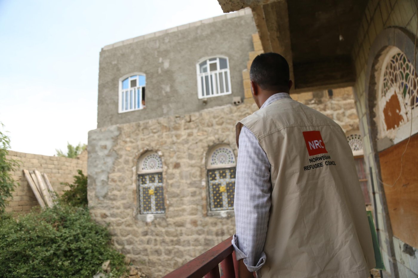 NRC staff member standing on a balcony and looking over the railing. Houses stand in the background. Staff member is wearing an NRC vest and the logo is clearly shown to the camera.