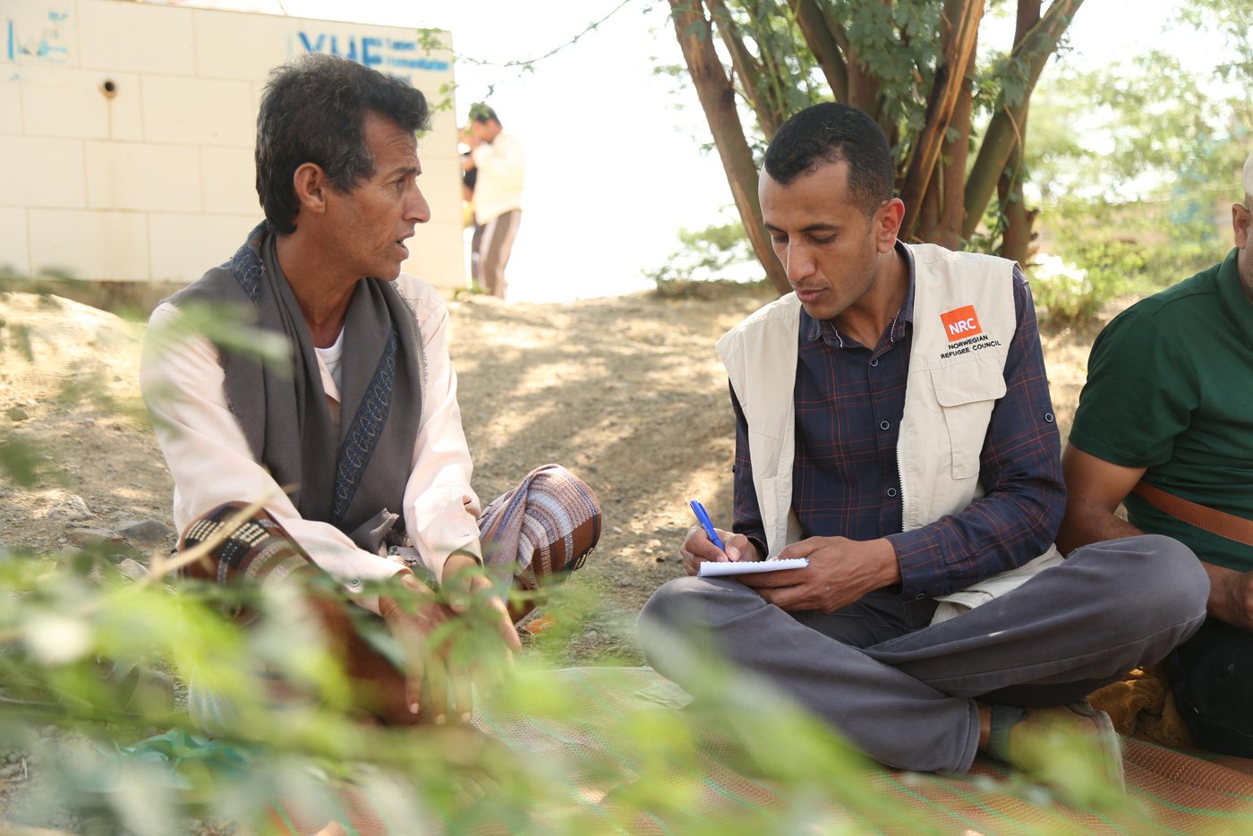 Landscape shot of Faisal talking to an NRC staff member. They are both sitting cross-legged on the ground amidst trees. The NRC staff member is writing notes in a notepad and is wearing an NRC branded vest.