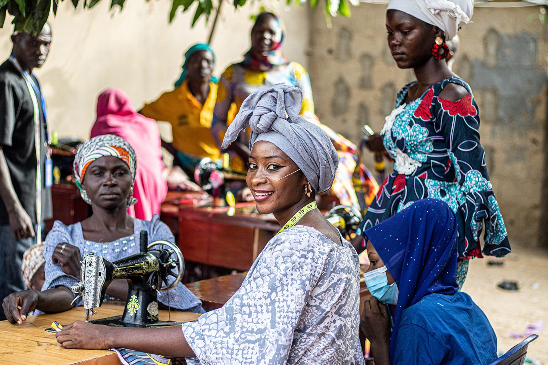 A woman is sitting by a sewing machine, teaching sewing to several other women. She is turned towards the camera, smiling.