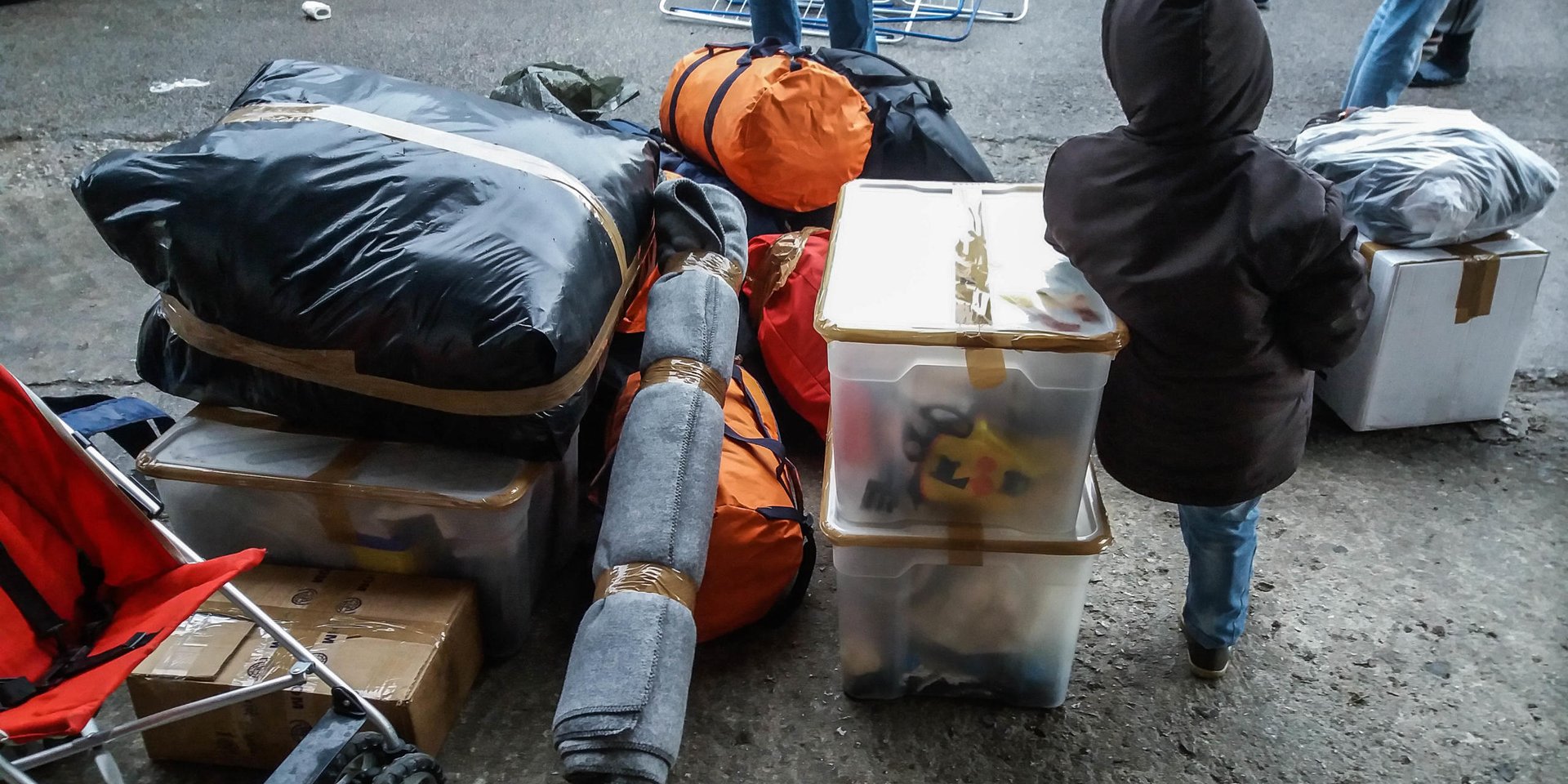 A small child stands next to packed and taped up boxes and bags, blankets and a stroller. 
