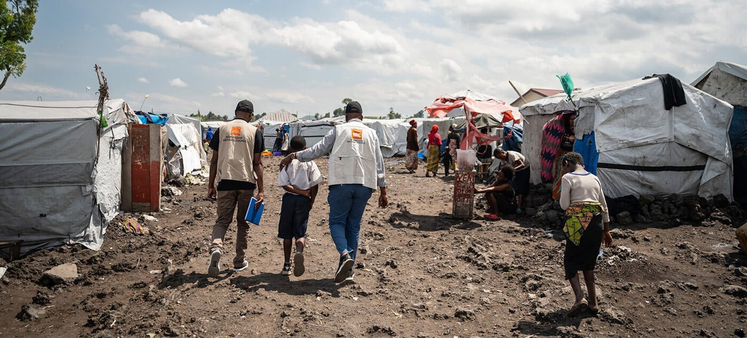 Two NRC staff walking with child through a row of tents in Goma, DR Congo.