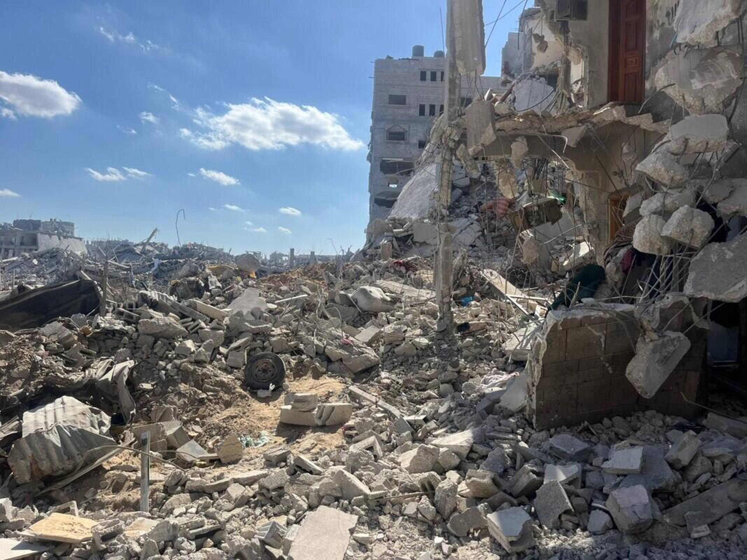 Sönderbombade byggnader och stora mängder bråte i ett bostadsområde i Gaza, med blå himmel i bakgrunden.