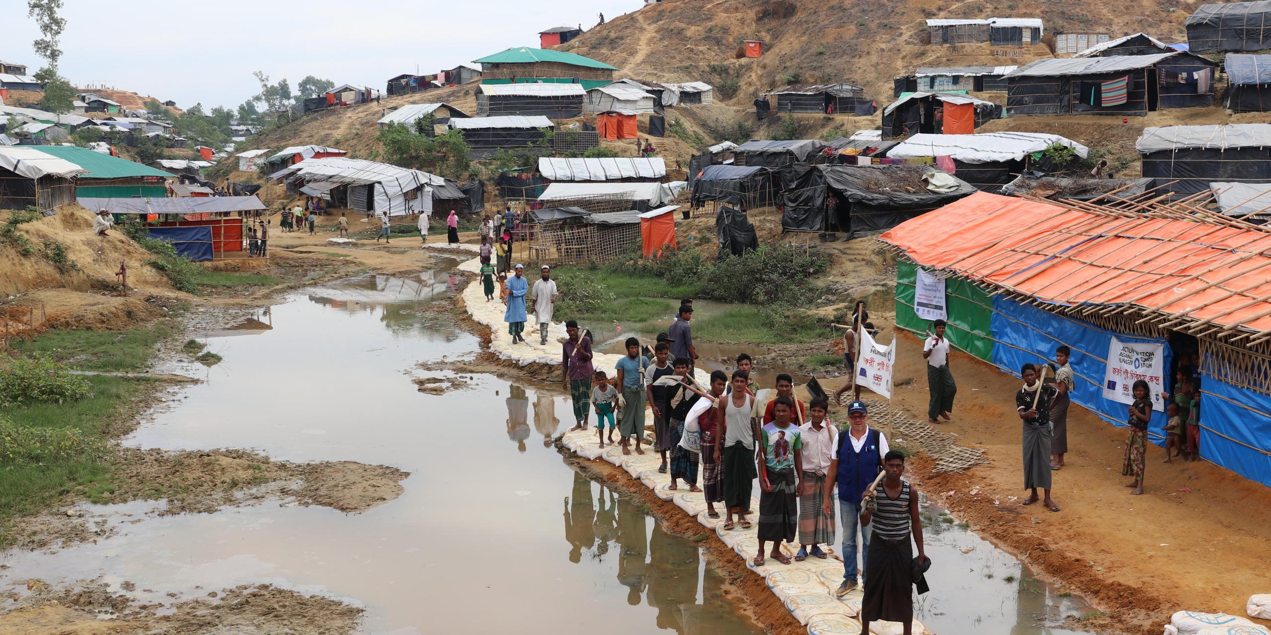 Small shacks on a hill behind a bridge where people walk over a swamp area in the Cox's Bazar camp.