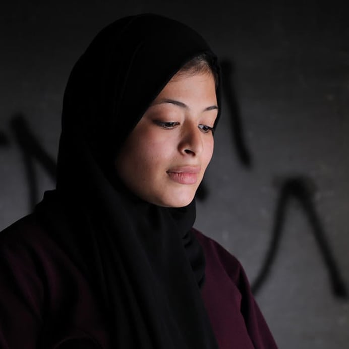 Portrait of Omaia, a 16-year-old Palestinian girl. She is standing in front a grey wall, looking down.