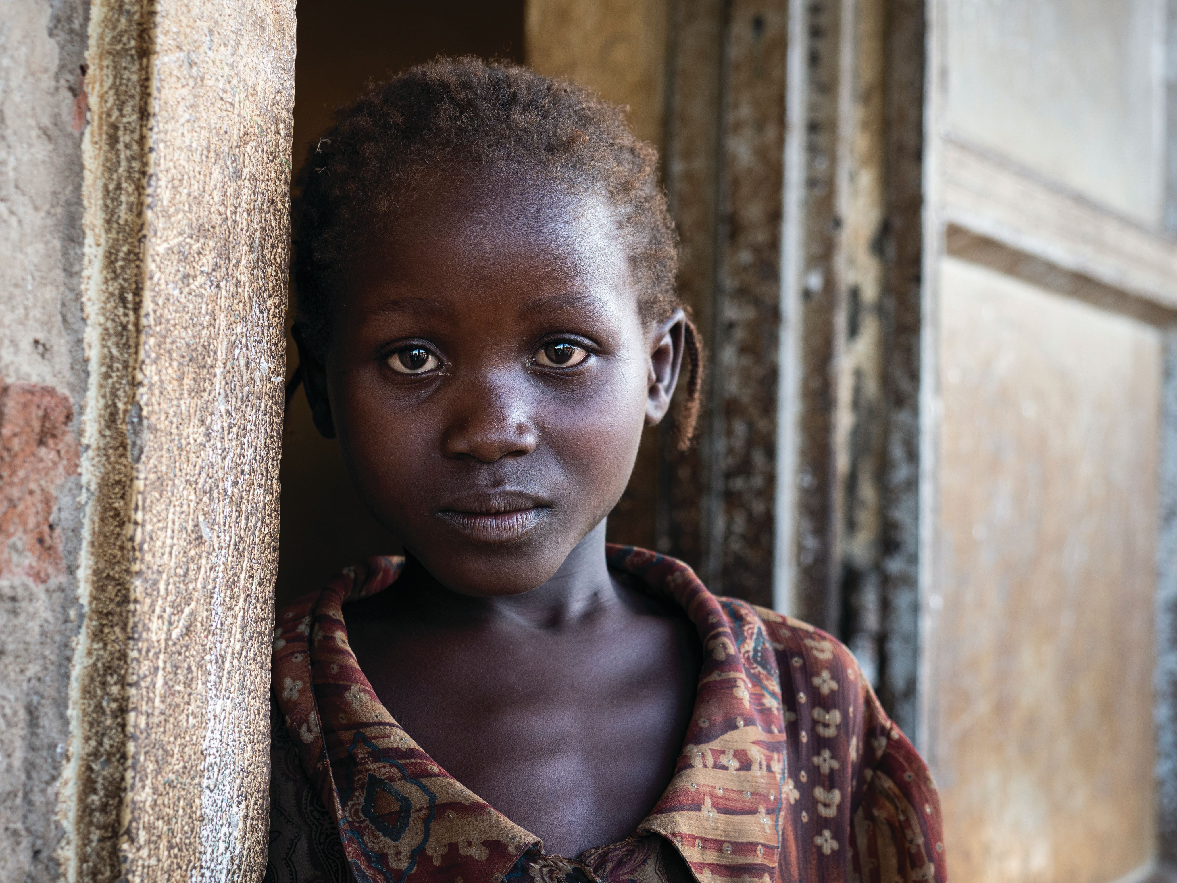 A 4-year-old South Sudanese girl leaning against a door frame, looking sombrely at the photographer.