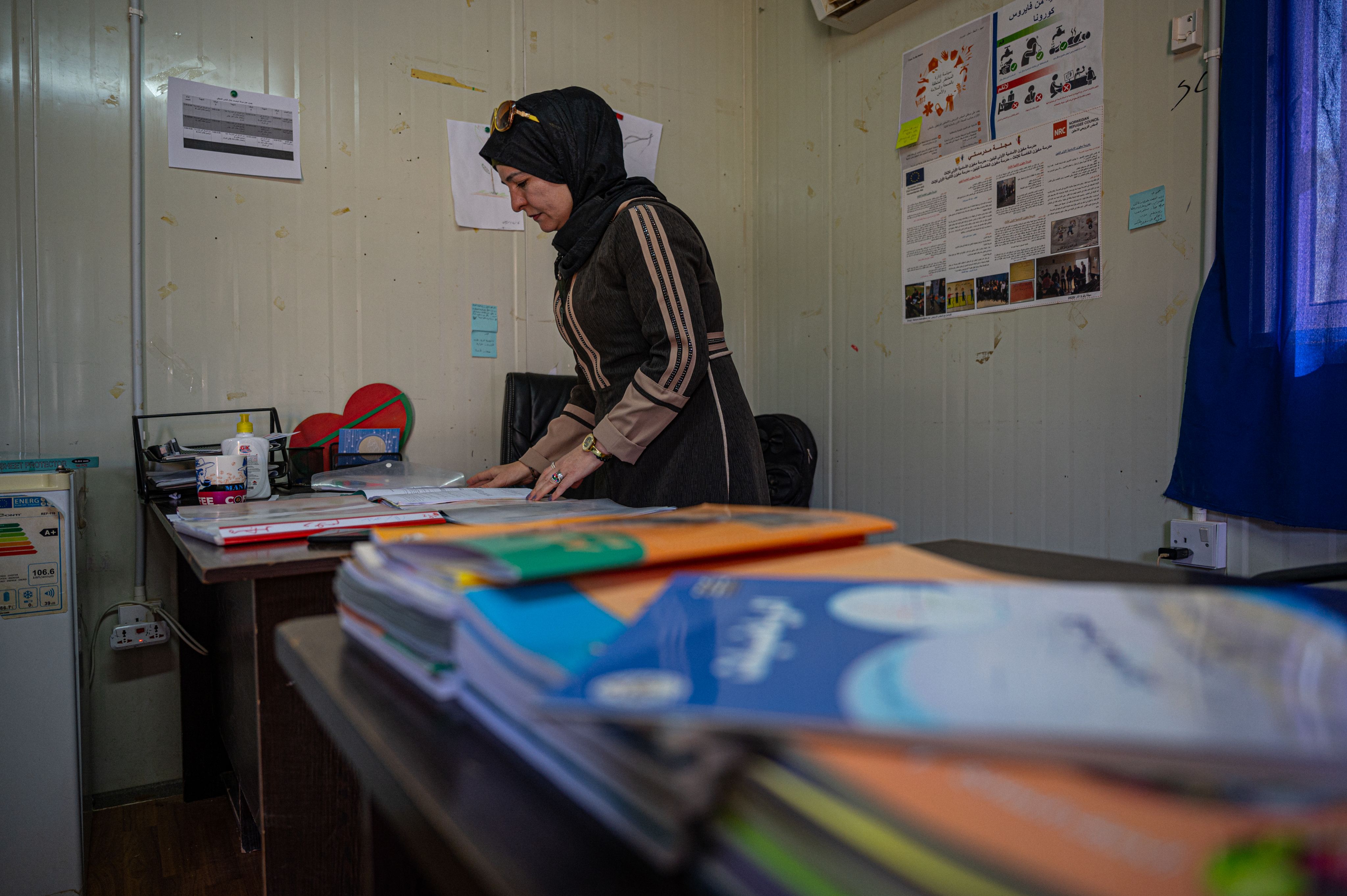 A Syrian teacher stands at her desk and looks through some papers