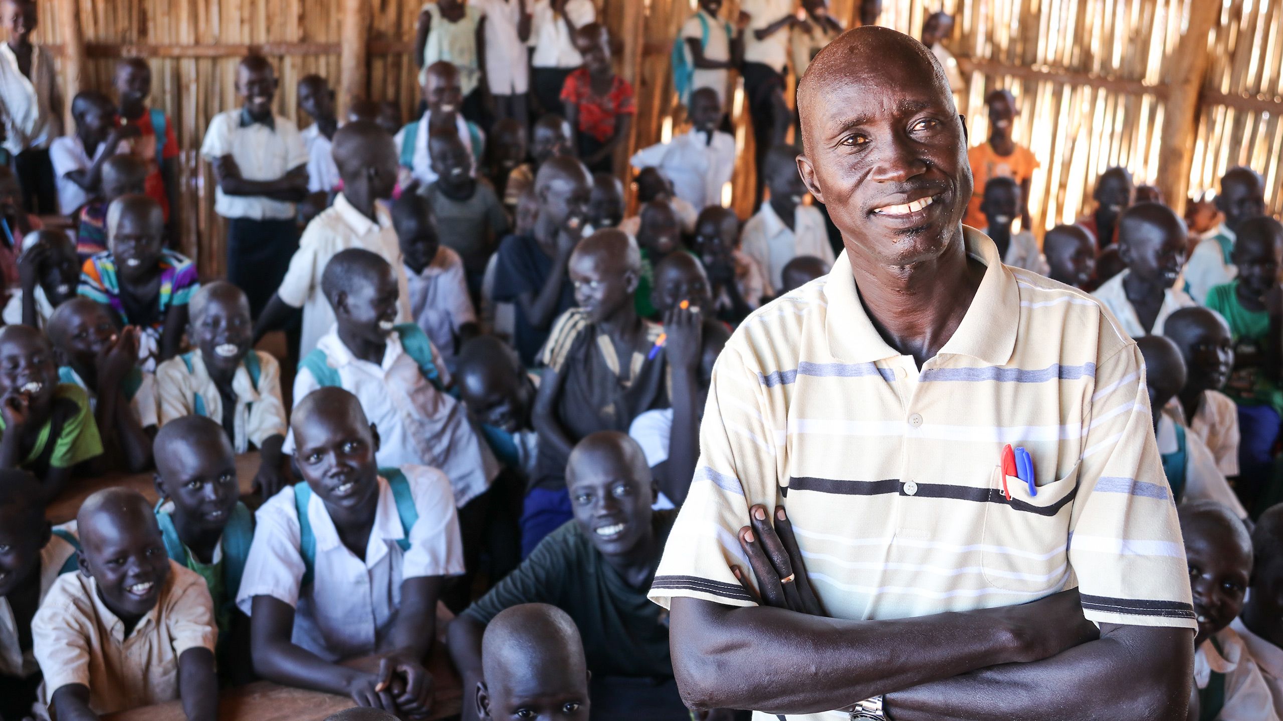 A man, with his armes crossed, smiling to the camera. Behind him is classroom full of students.