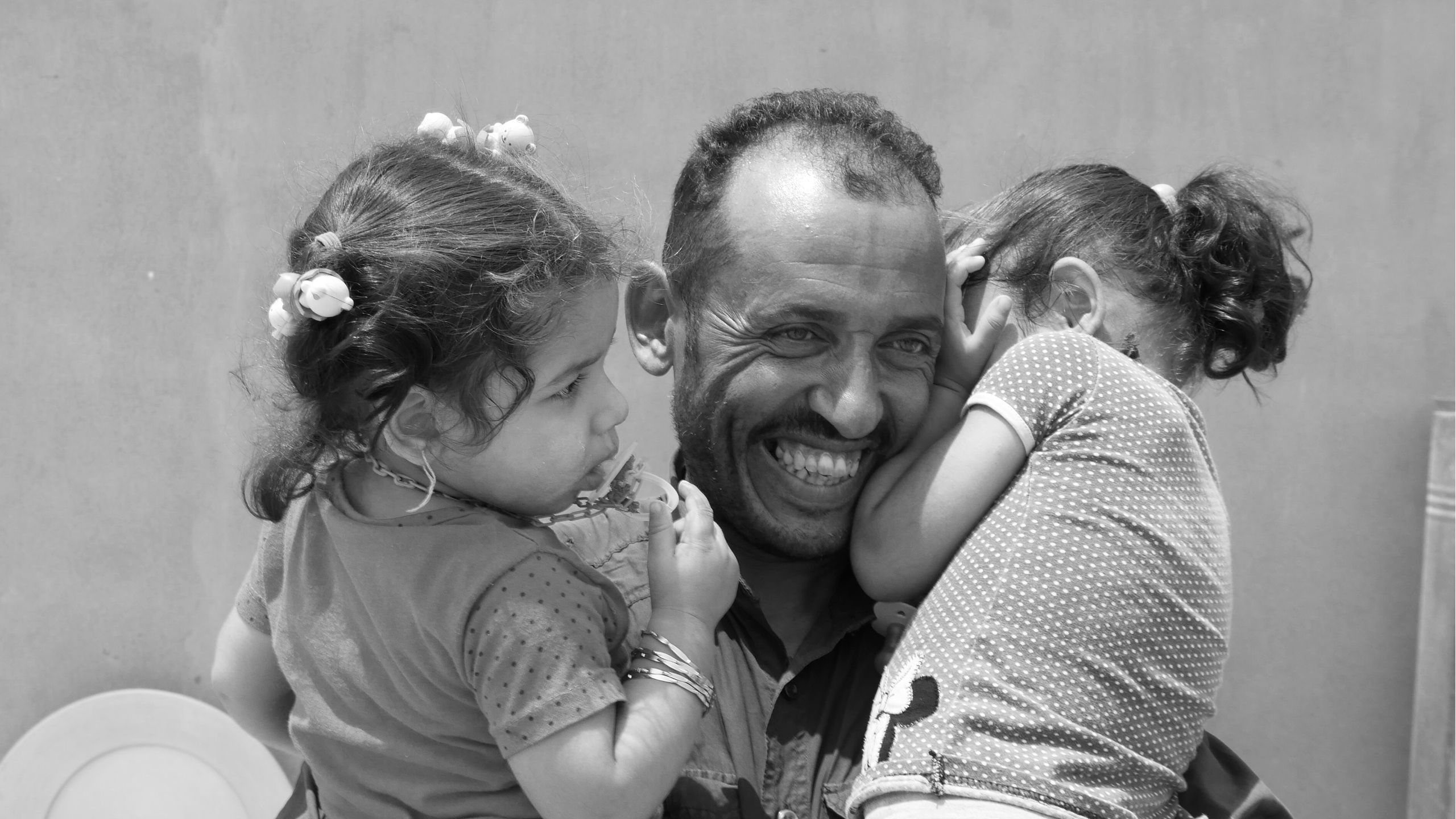 A man smiling wide, carrying his two daughters 