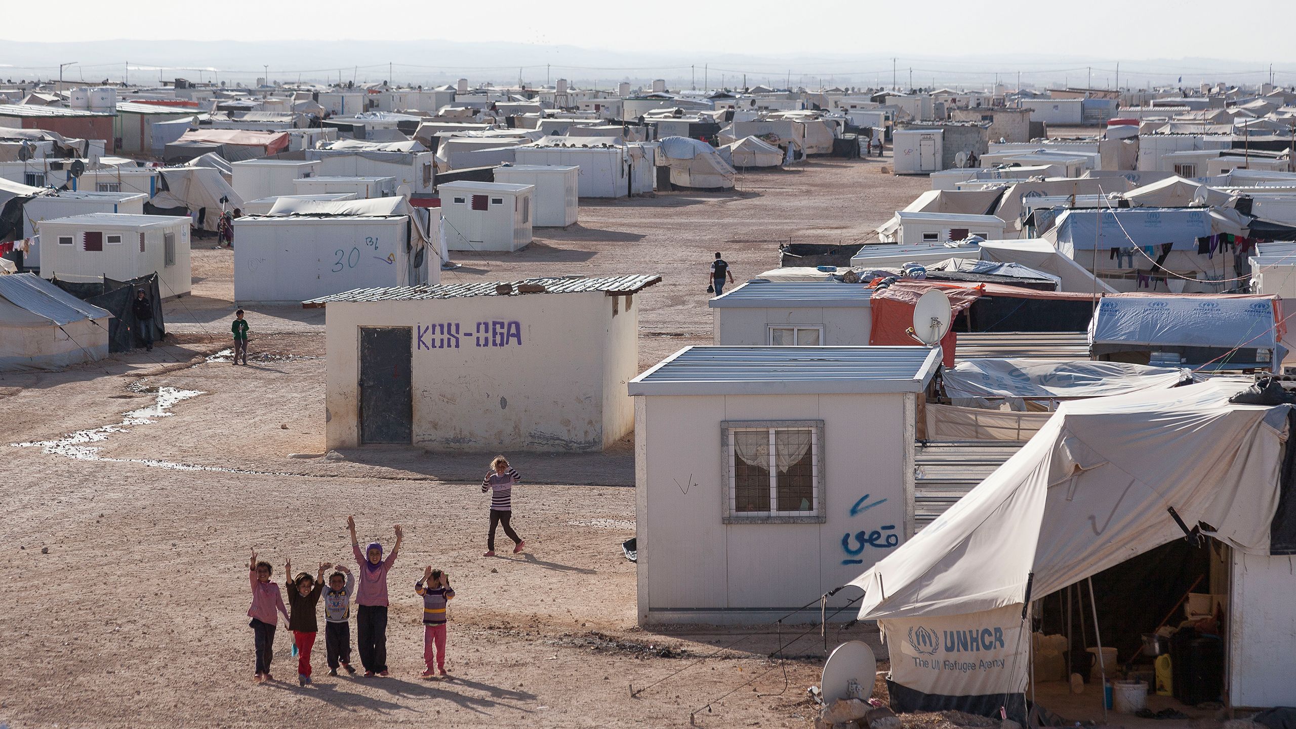 Fem jenter står ved siden av hverandre og vinker til fotografen. Bak dem strekker Zaatari-leiren i Jordan seg utover.