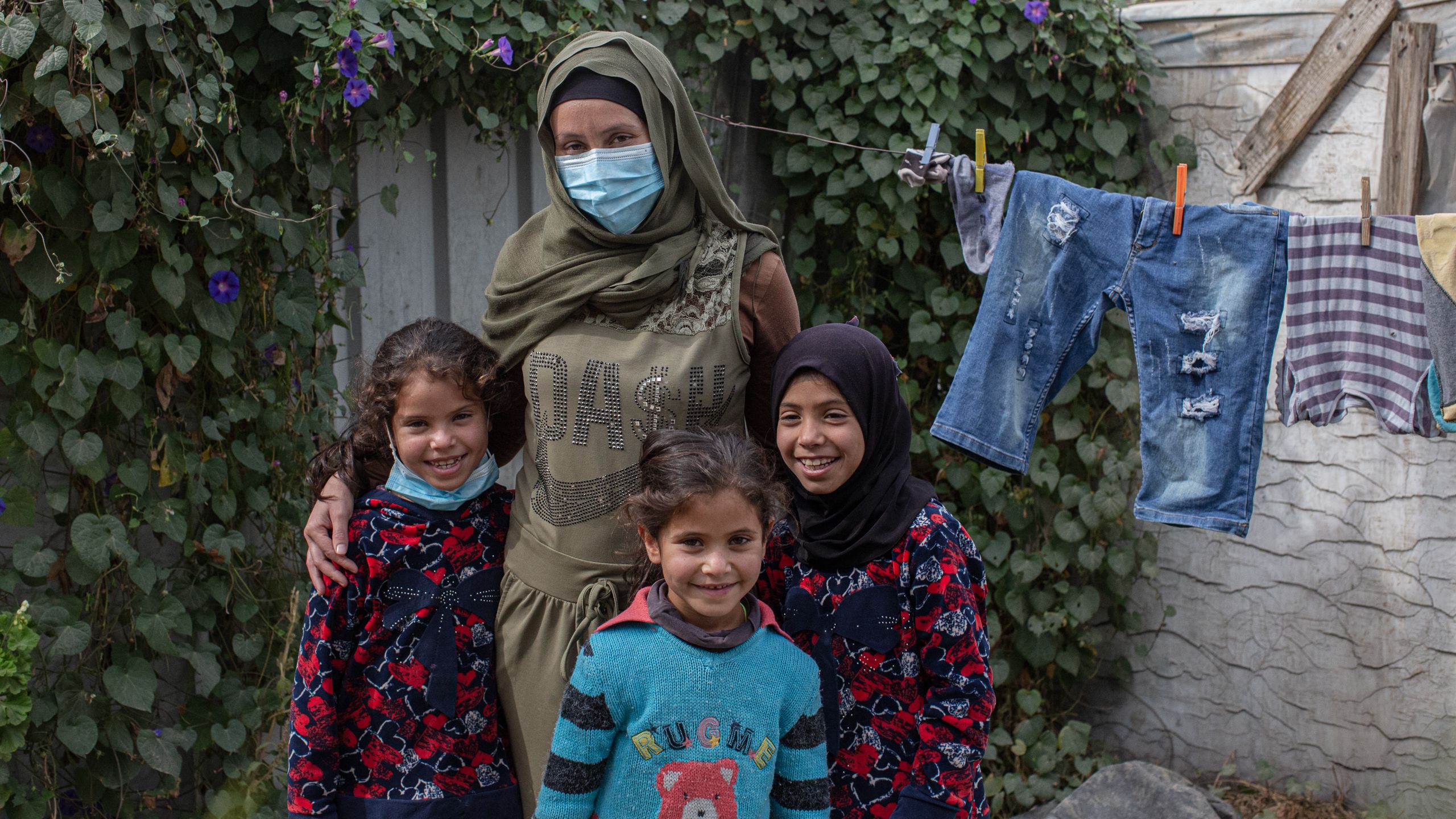 En kvinne og hennes tre døtre står utenfor teltet sitt. En snor med klesvask går bak dem. På veggene vokser det tett med klatreplanter.