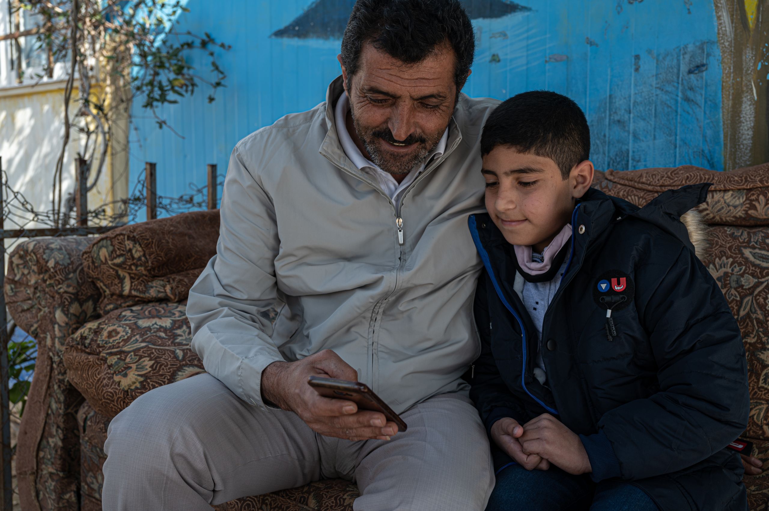 Osama, 9, and his father, Mohammad, 49, sit on a couch. Mohammad is showing Osama something on his phone.
