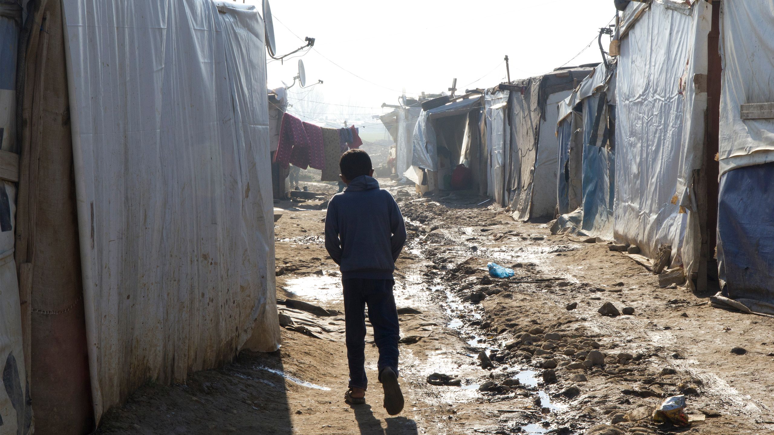 A young boy treading the muddy ground between tents in a refugee camp.