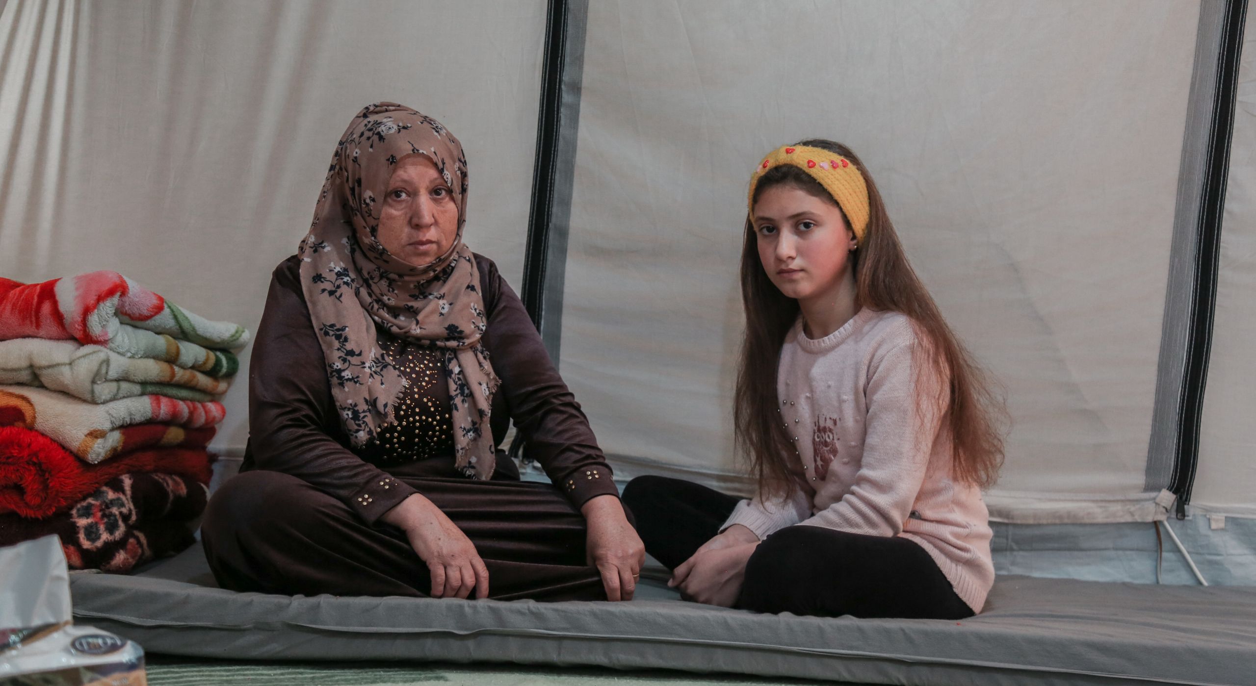 Feryal, 50, and her daughter Noor, 12, sitting on a grey mattress in their tent.