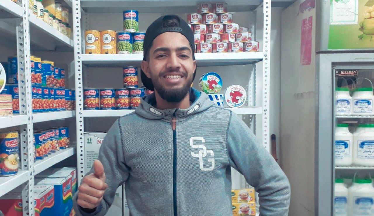 Portrait of Wassim in front the shelves in his shop. He is smiling and showing thumbs up.